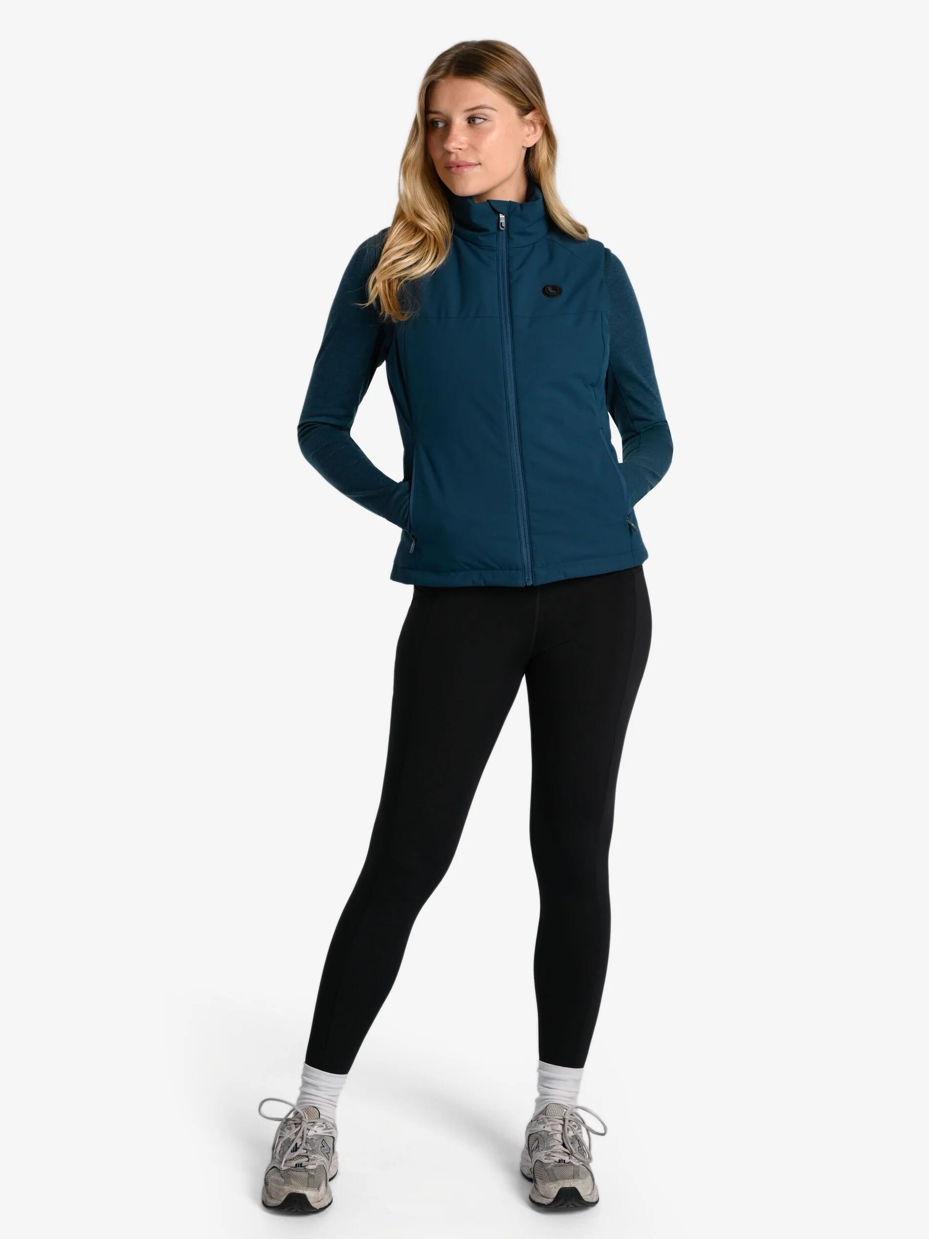 A person wearing a blue zip-up jacket, black leggings, and gray sneakers stands with hands in pockets against a plain white background.