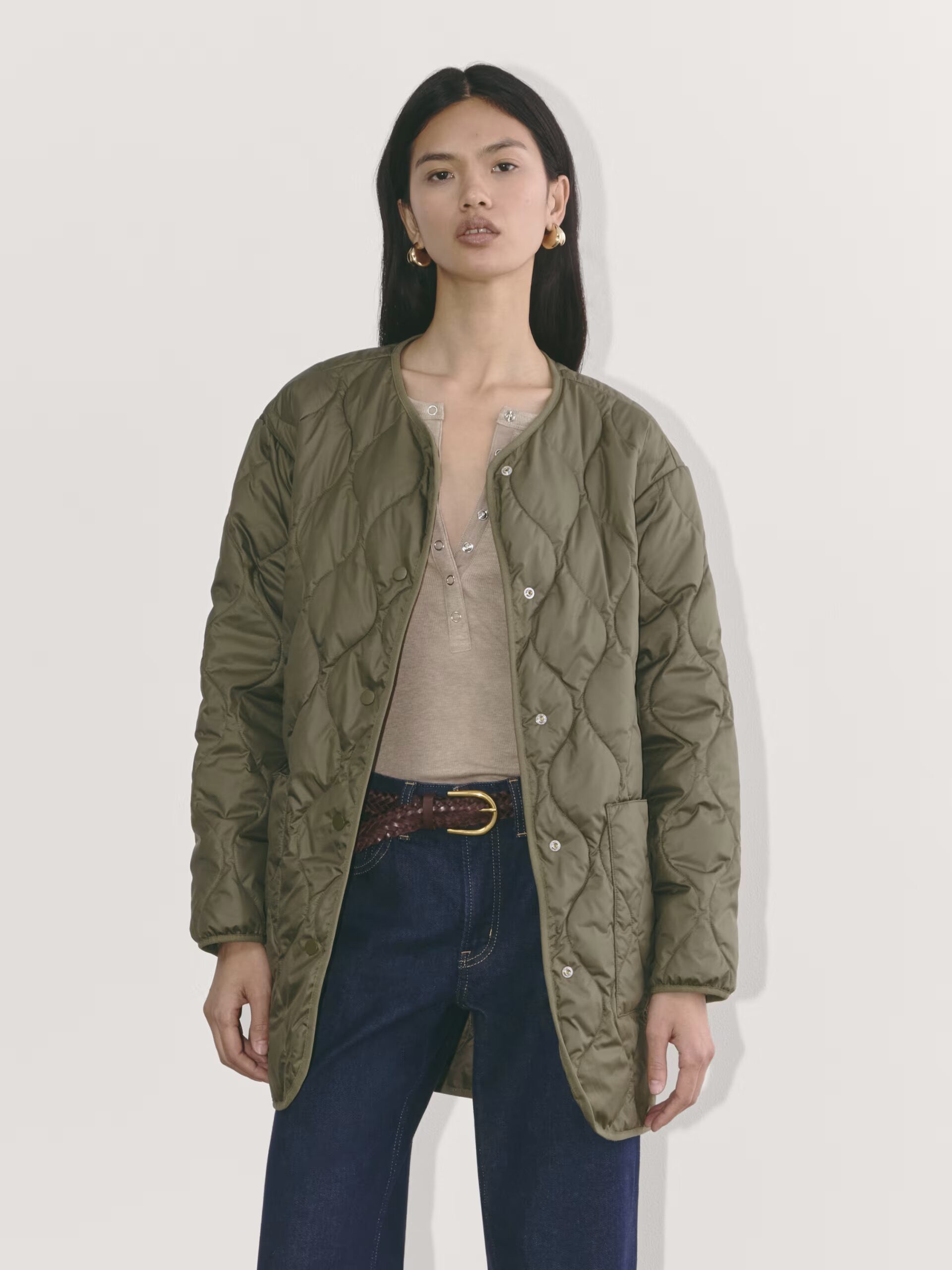 A woman wearing a quilted green jacket over a beige top and dark jeans stands against a plain background.