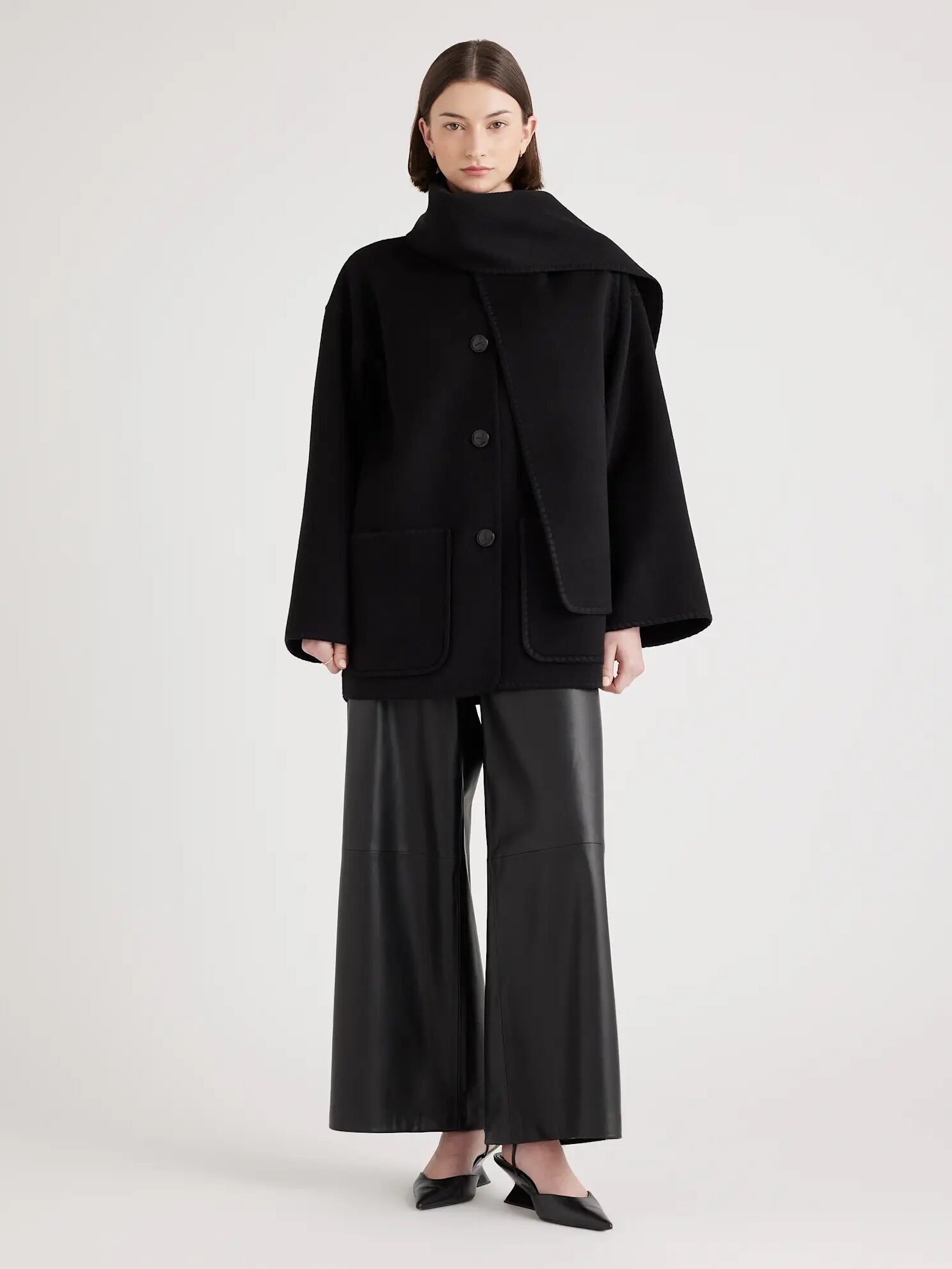 Person wearing a black coat with large buttons, leather wide-leg pants, and pointed shoes, standing against a plain background.