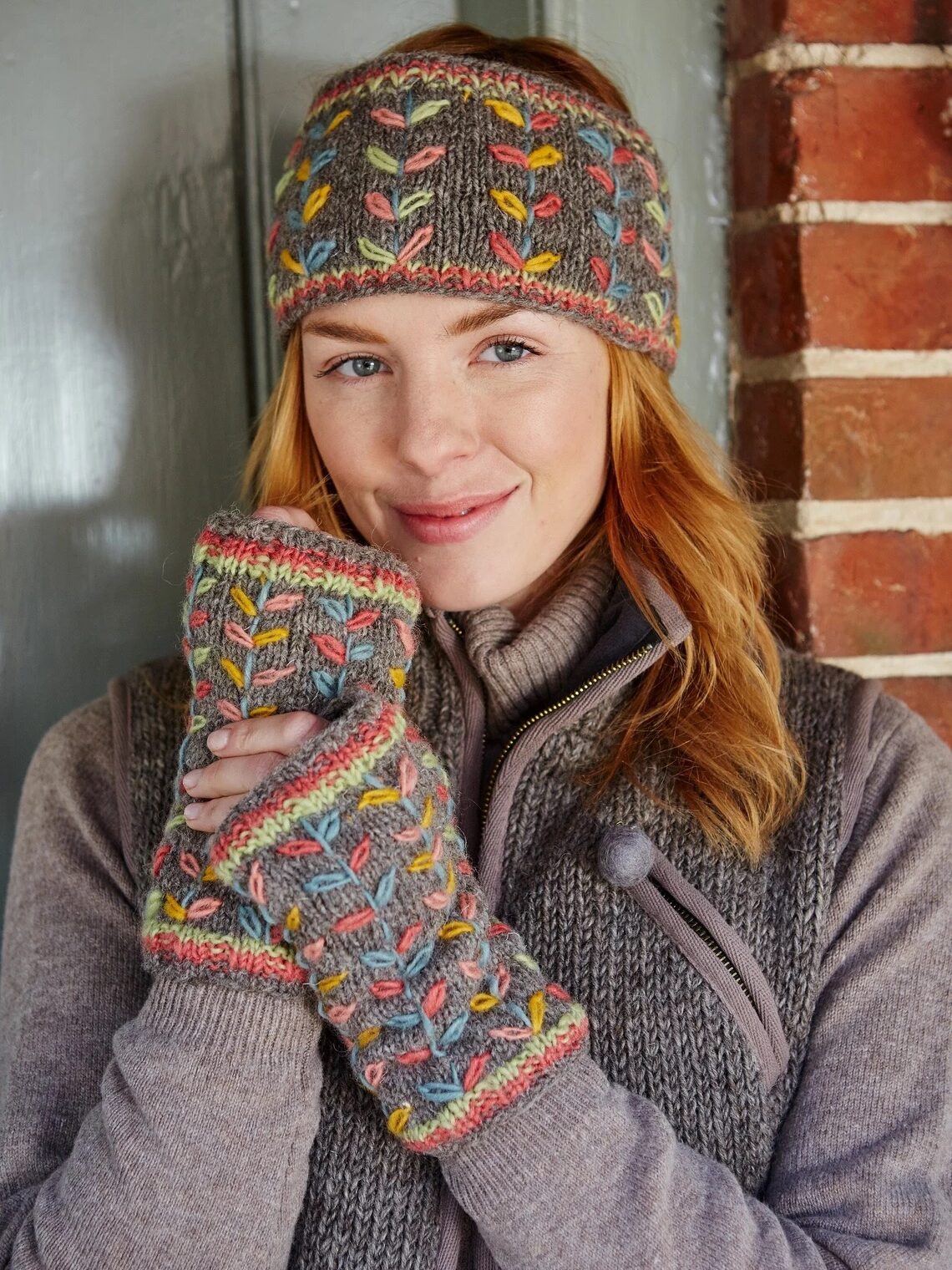 Person wearing a knitted headband and fingerless gloves, both with a colorful pattern. They are standing in front of a brick wall, dressed in a sweater and vest.
