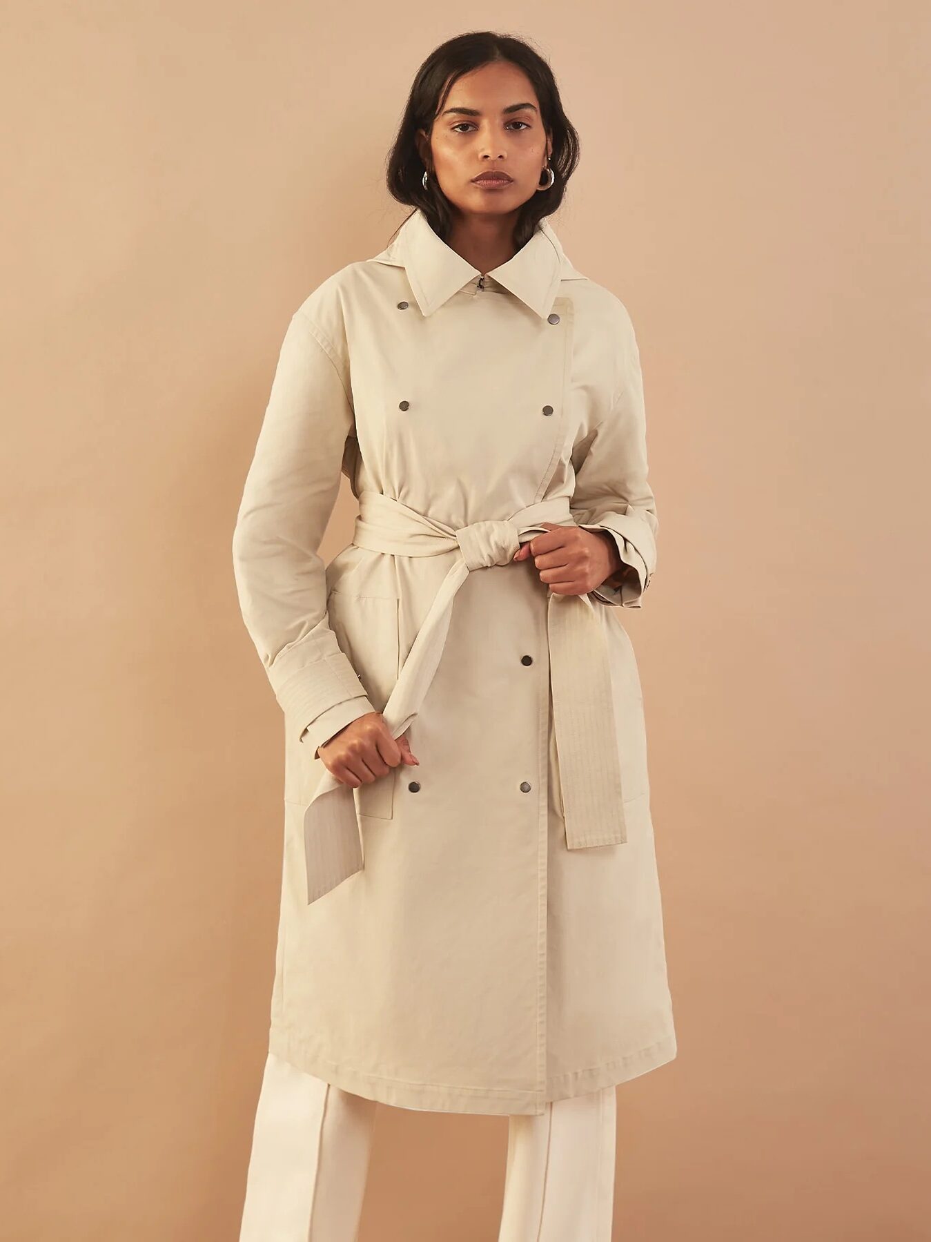 Person wearing a beige trench coat with a tied belt and standing against a plain background.