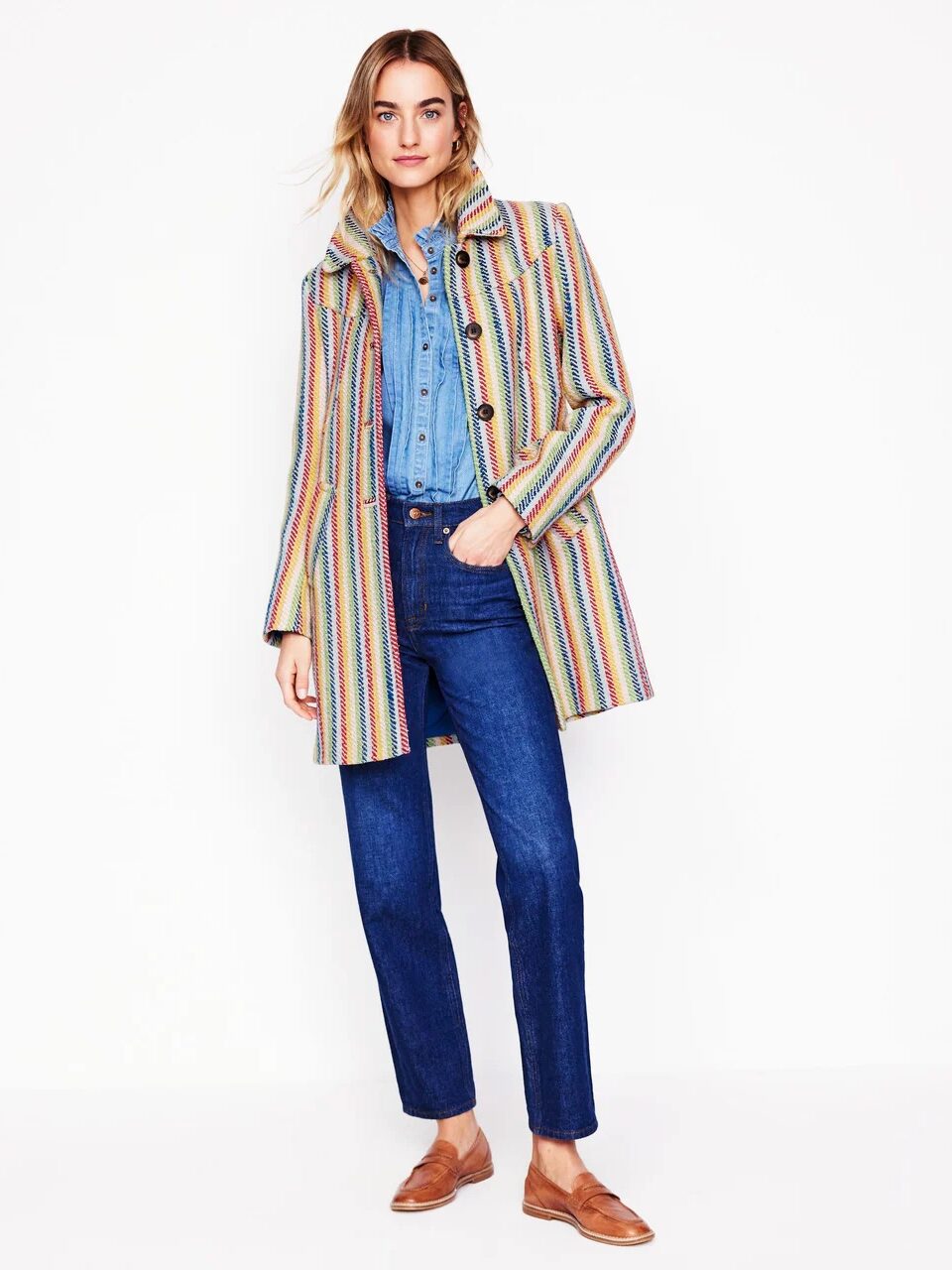 Person wearing a multicolored striped coat, denim shirt, blue jeans, and brown shoes stands against a plain white background.