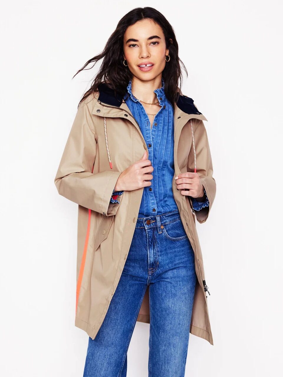 Woman wearing a beige hooded coat over a blue denim shirt and jeans, standing against a white background.
