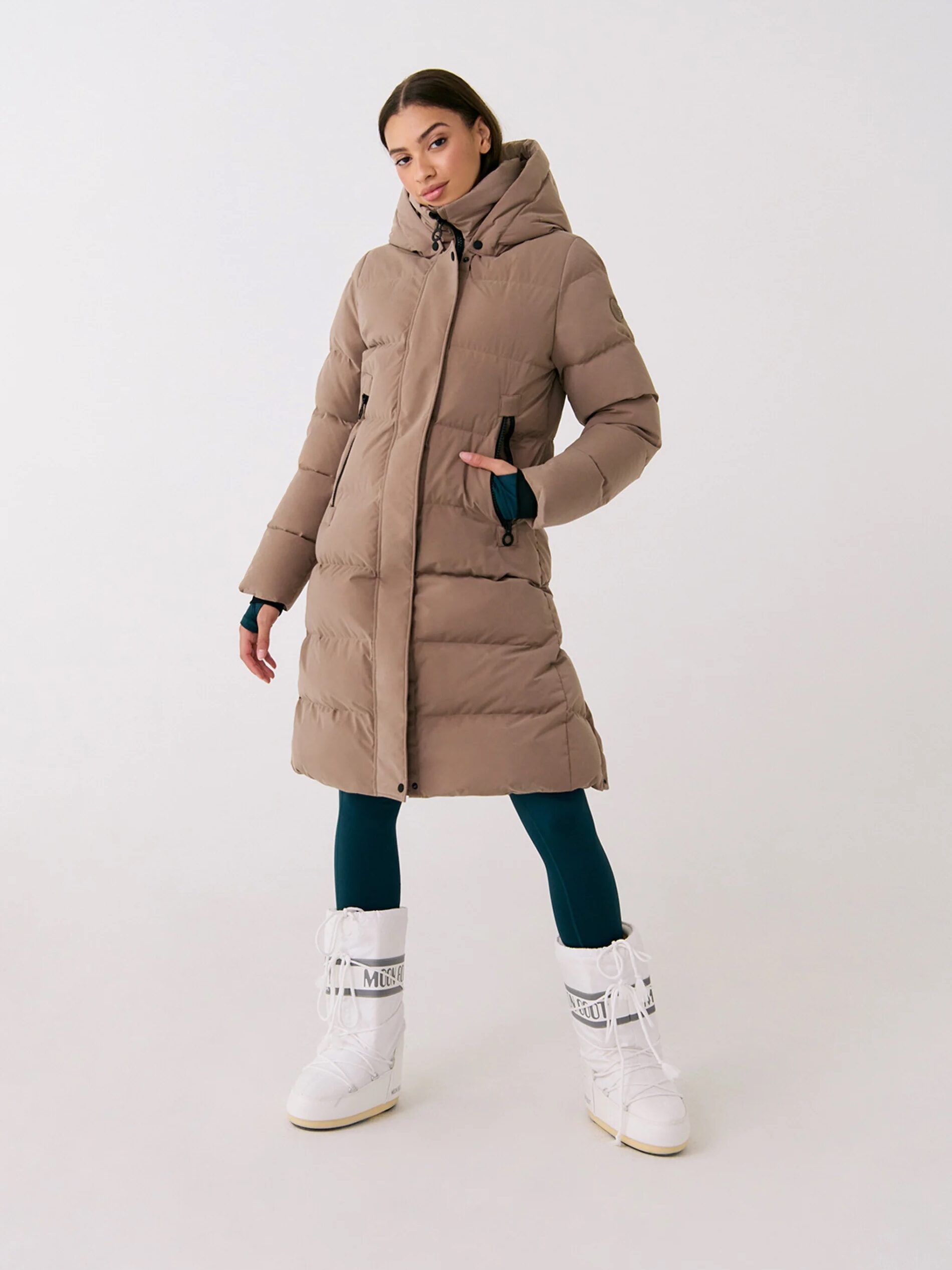 Person wearing a long brown puffer coat, dark leggings, and white snow boots, standing against a neutral background.