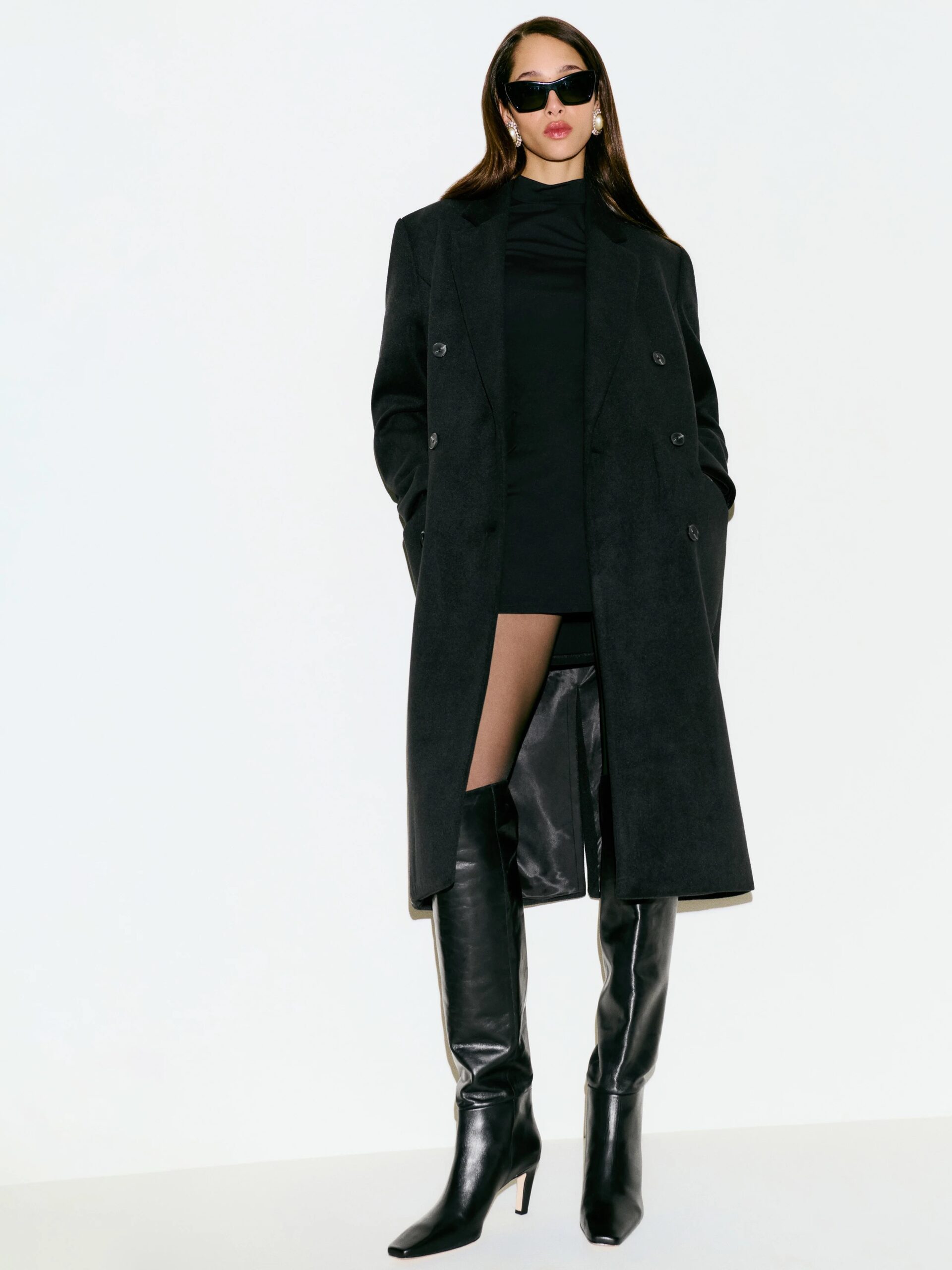 Person wearing a long black coat, black dress, and tall black boots, standing against a plain background.