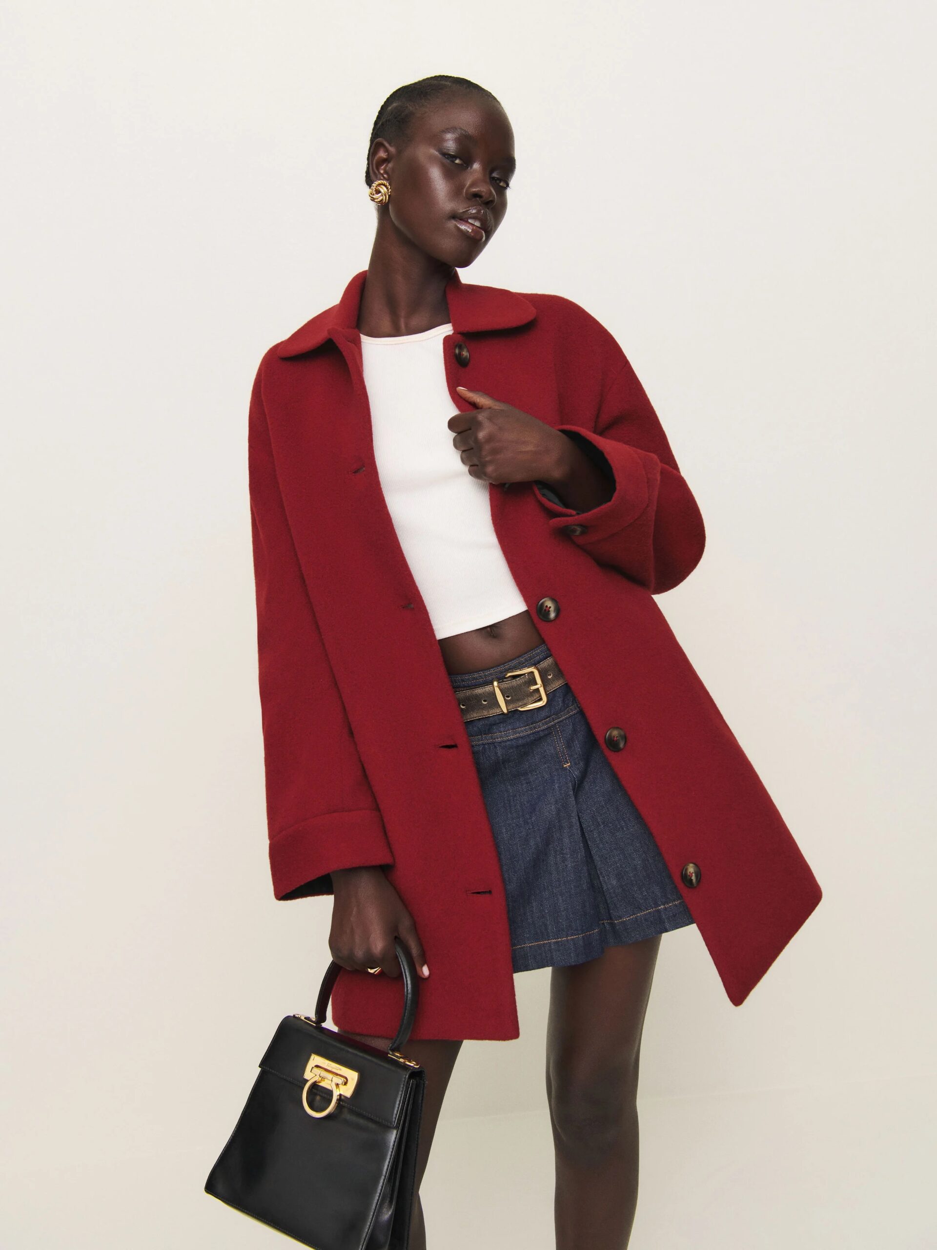 A person poses in a red coat, white top, and denim skirt, holding a black handbag.