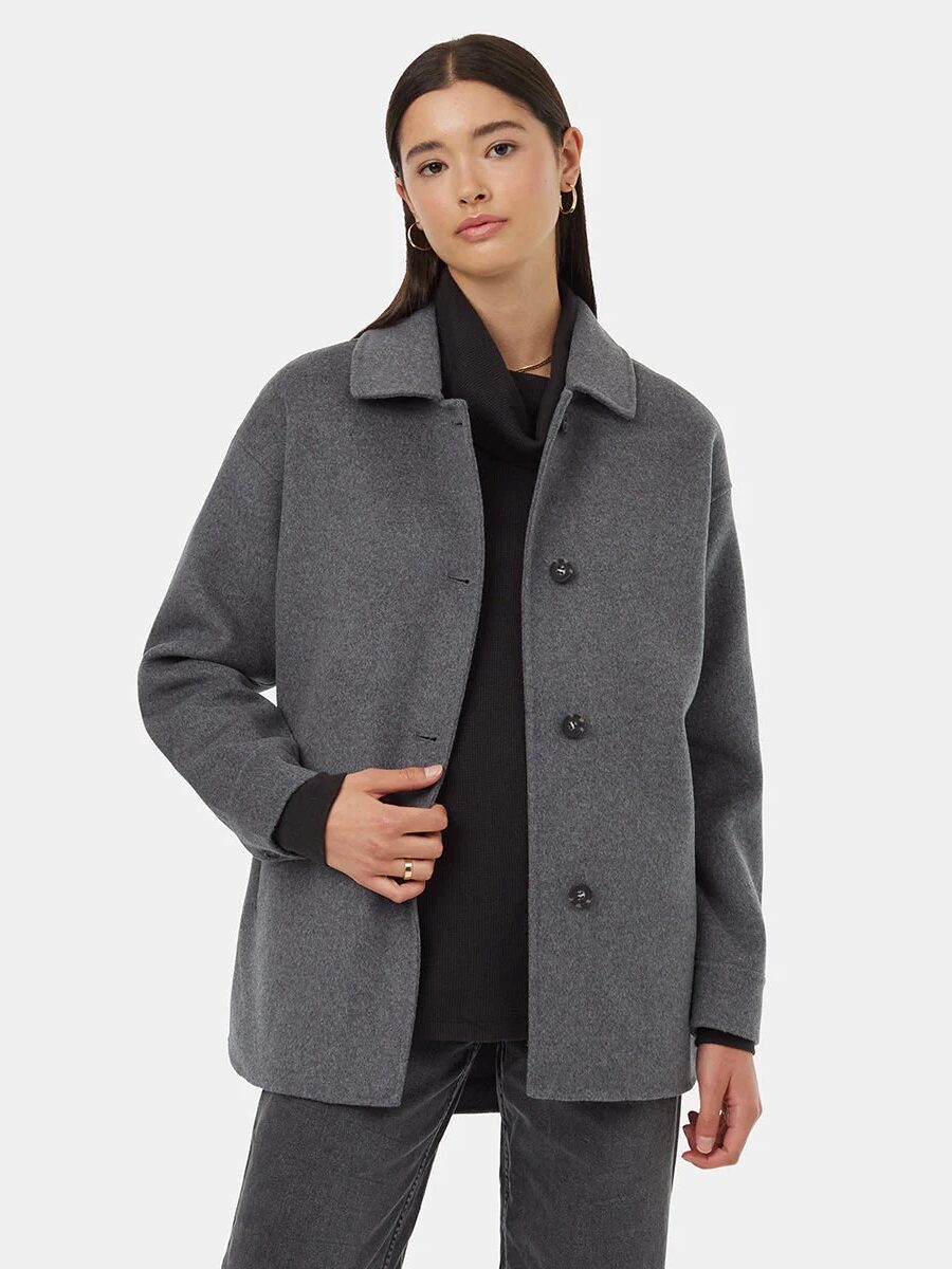 A person wearing a gray button-up coat over a black top and dark jeans stands against a plain white background.