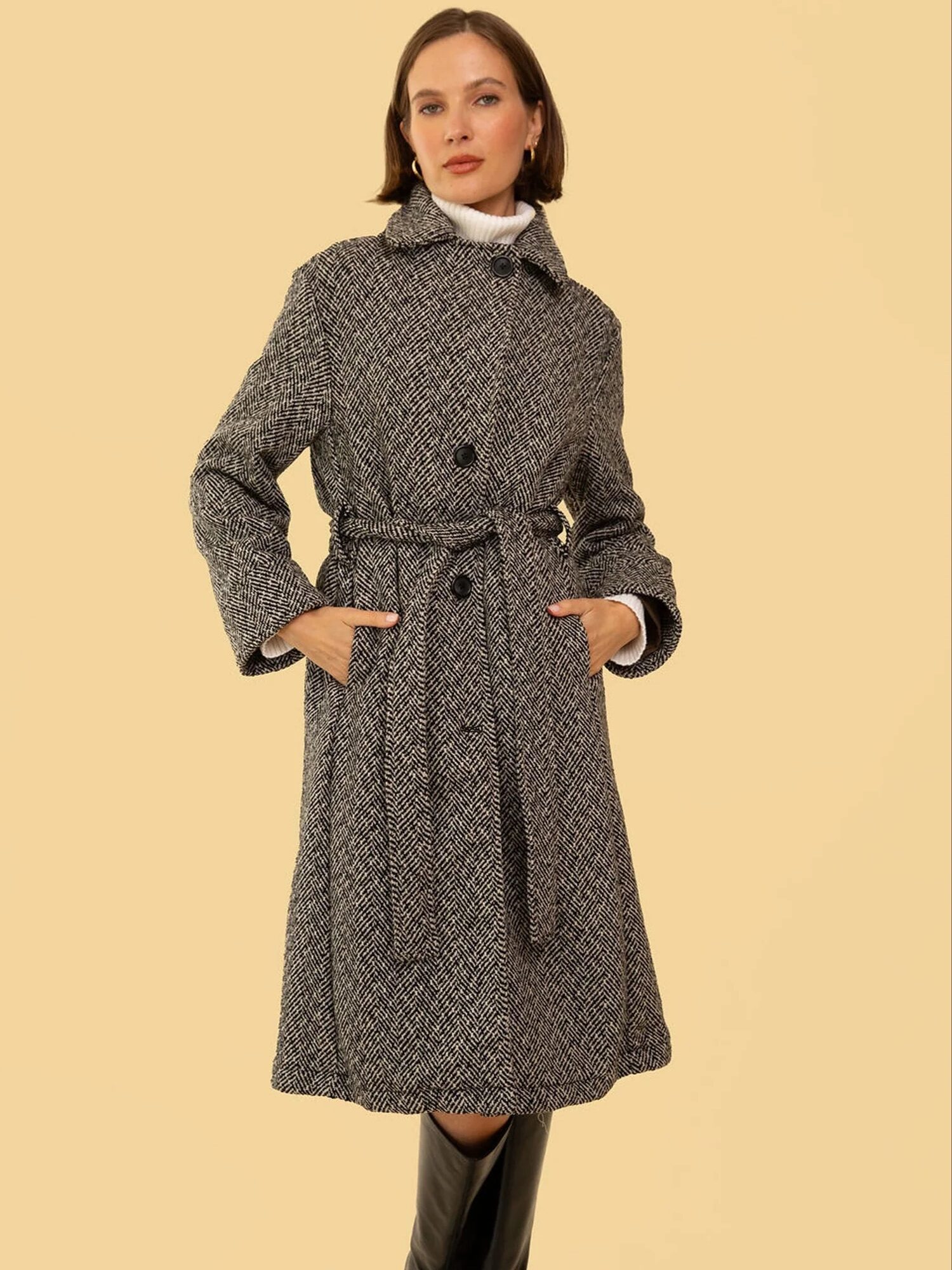 Person wearing a gray herringbone trench coat with a belt, hands in pockets, and black boots, standing against a beige background.