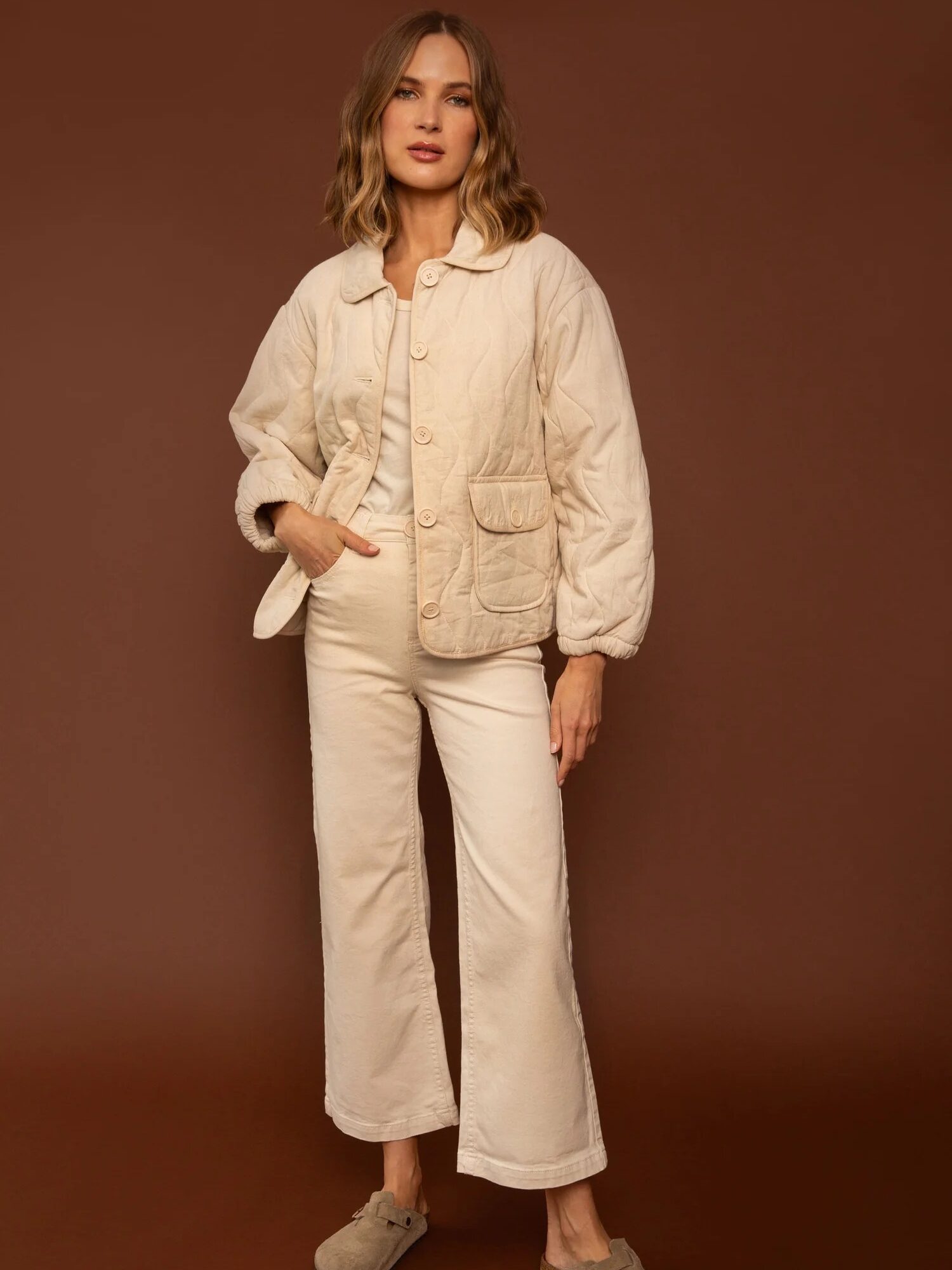 A person wearing a beige, quilted jacket and pants stands against a brown background.