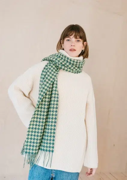 Person wearing a white sweater, blue jeans, and a green houndstooth scarf stands against a plain background.