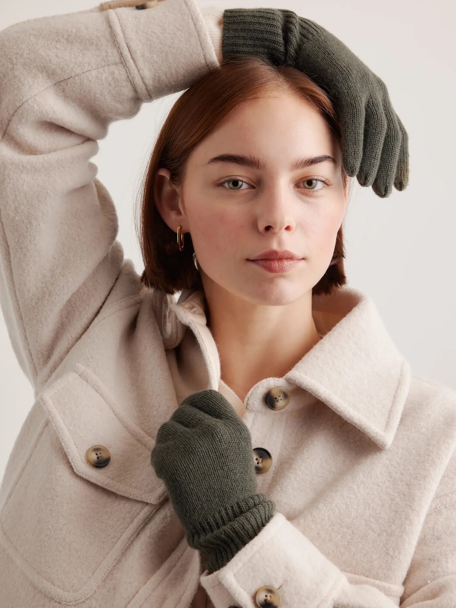 Person wearing a cream-colored coat and green gloves poses with one hand on head and the other on chest, looking at the camera.