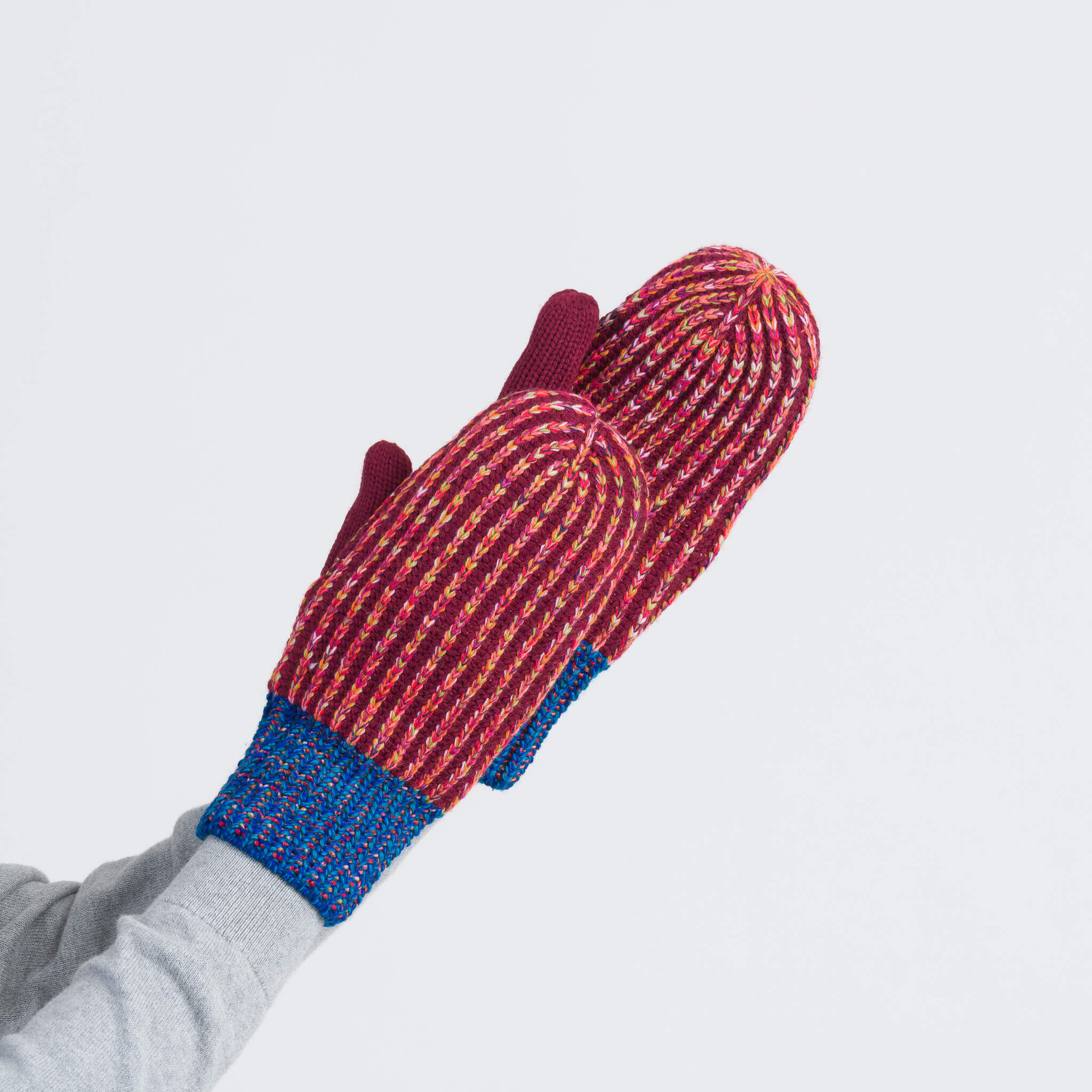 A person wearing colorful knitted mittens with stripes and blue cuffs raises their hands against a plain white background.