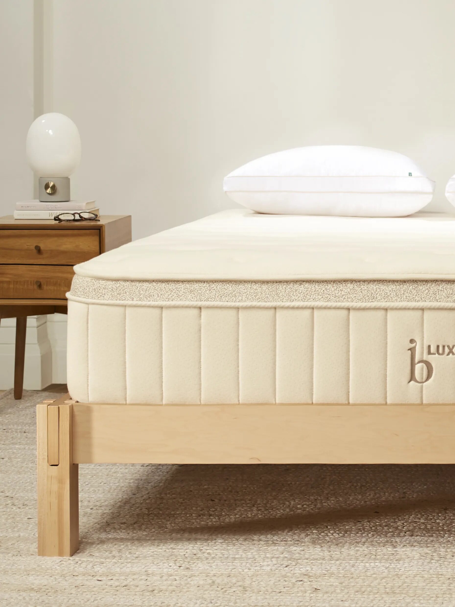 A beige mattress with "Luxe" logo on a wooden bed frame, next to a wooden nightstand with a lamp, book, and glasses.