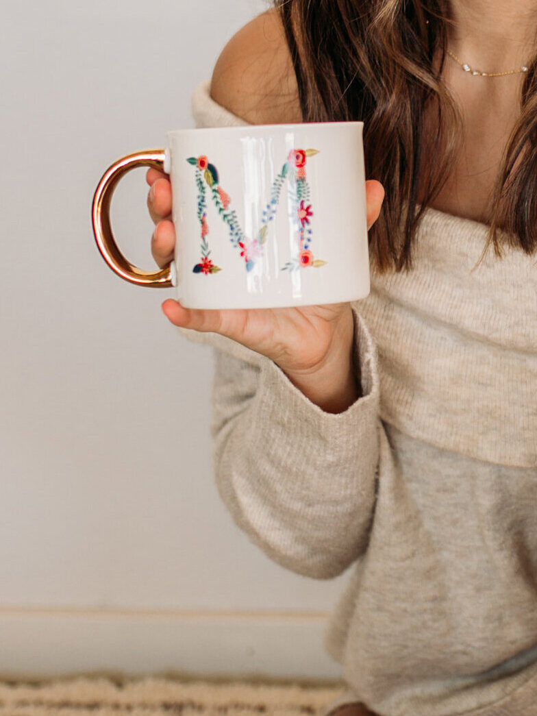 A person holding a white mug with a floral letter 'M' design and a gold handle, wearing a beige off-the-shoulder sweater.