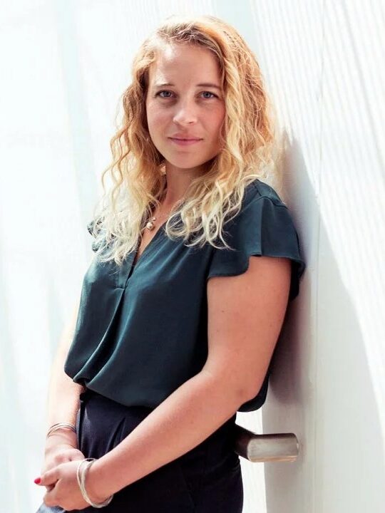 Woman with long curly hair leans against a wall in a well-lit, modern indoor space, wearing a dark blouse and pants, hands clasped.