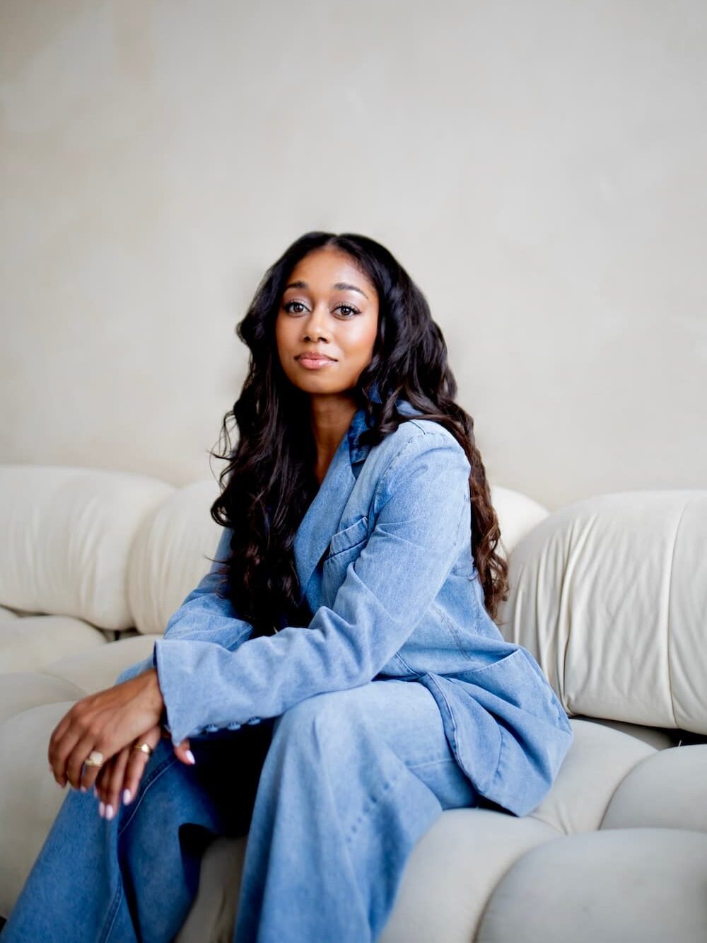 Woman with long hair wearing a light blue suit sits on a beige couch against a neutral background.