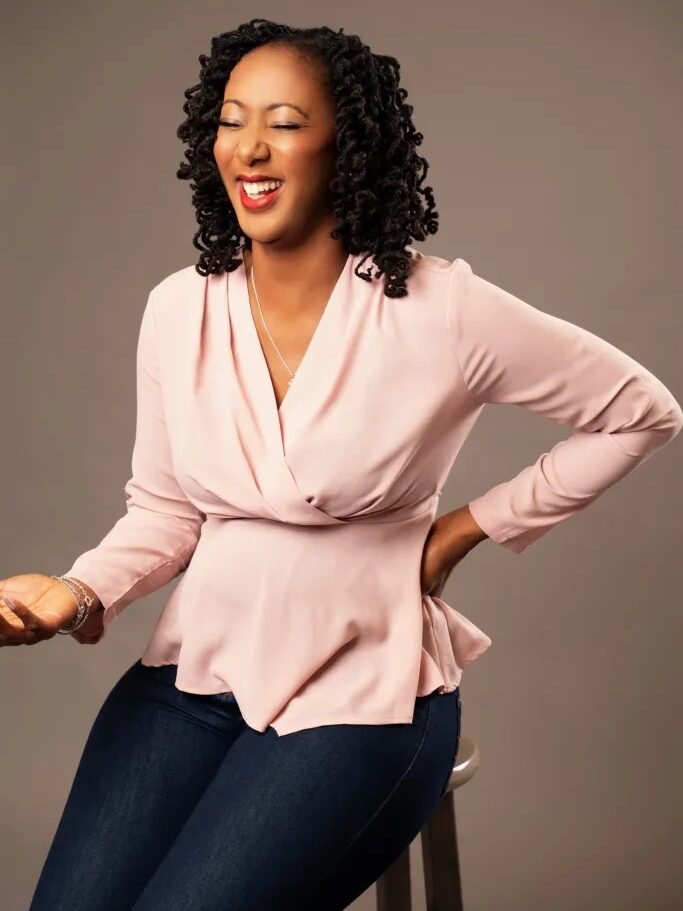 Smiling woman with curly hair, wearing a long-sleeve pink blouse and jeans, sits on a stool against a plain background.