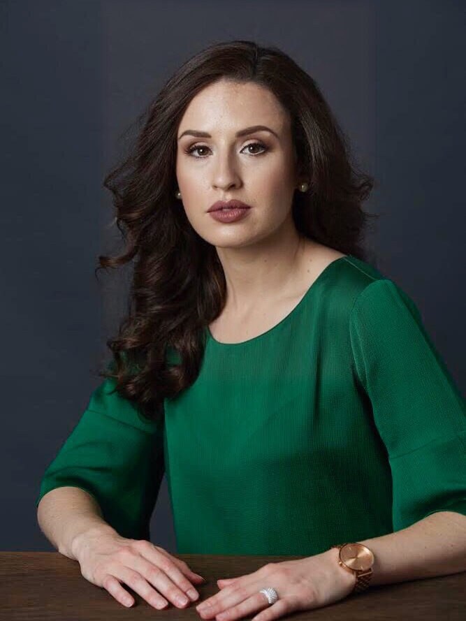 A woman with long dark hair in a green top and a watch sits at a wooden table against a dark background.