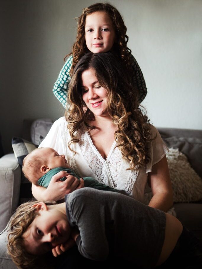 A woman sits on a couch with a baby in her arms, a child resting on her lap, and another child with arms on her shoulders, all looking relaxed.