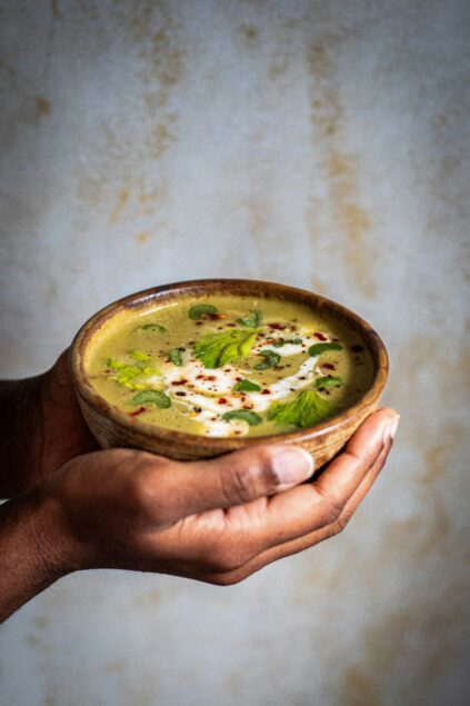 Hands holding a bowl of green soup, garnished with herbs, seeds, and a drizzle of white cream.