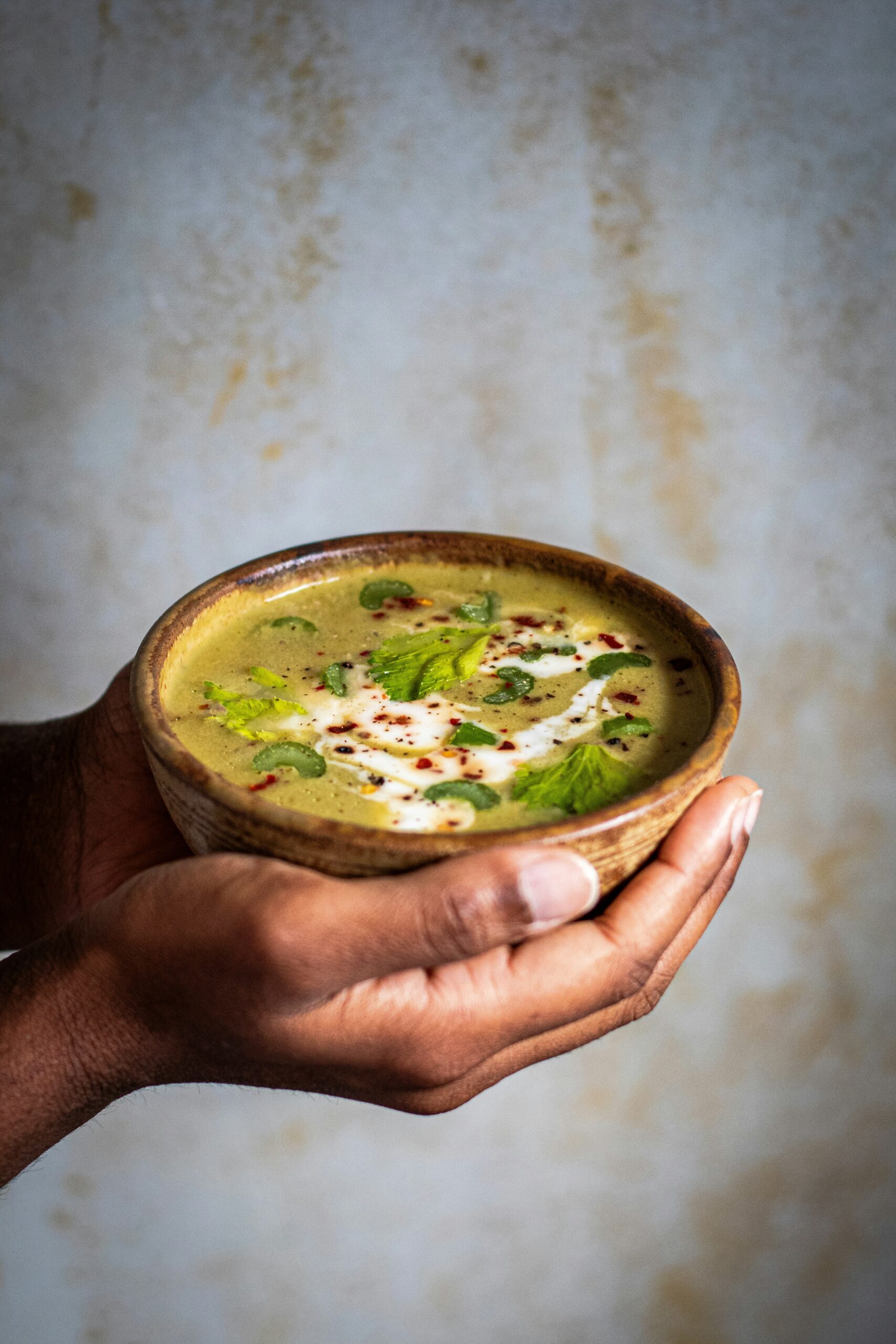 Hands holding a bowl of green soup, garnished with herbs, seeds, and a drizzle of white cream.