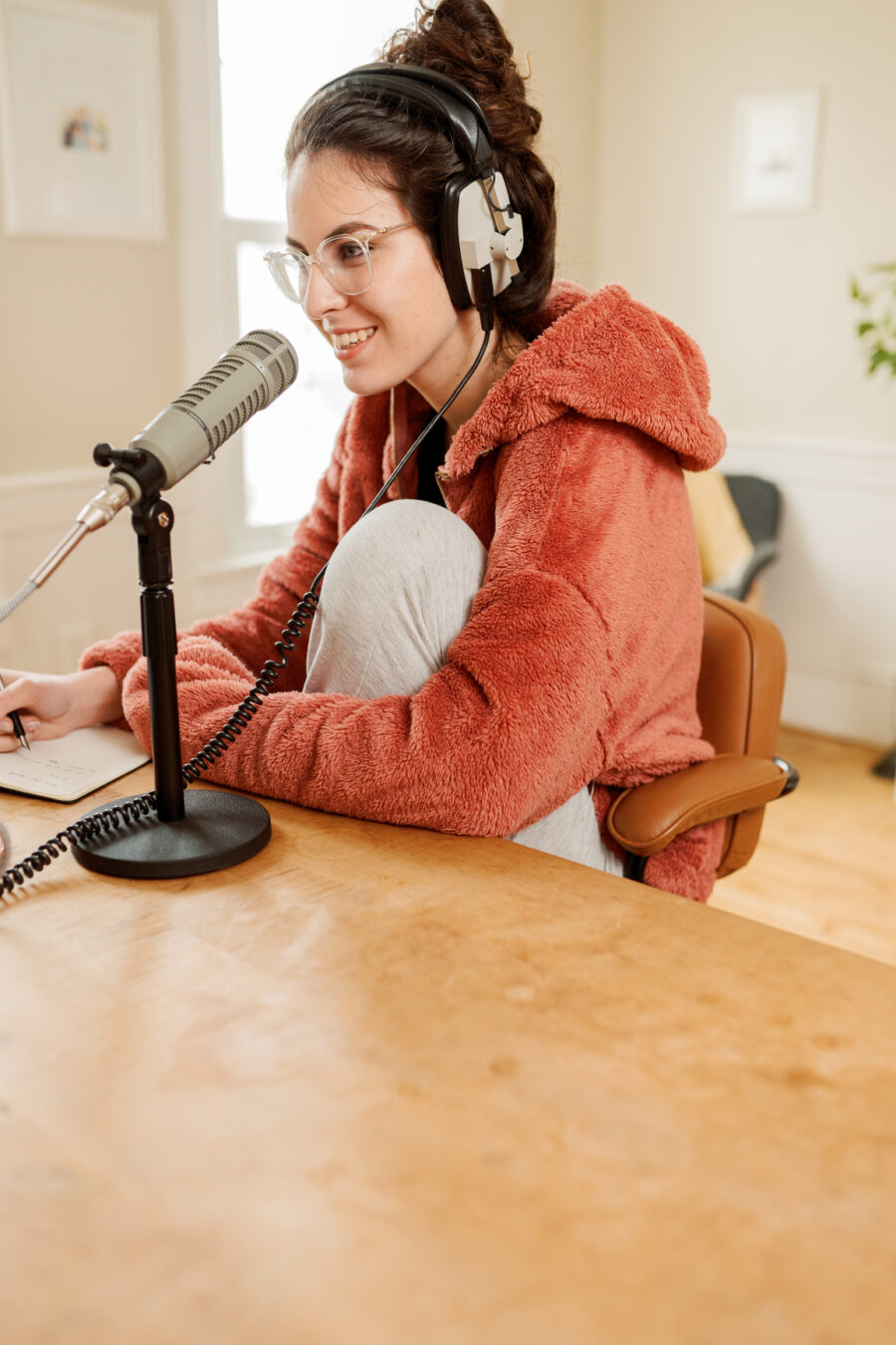 Person wearing headphones and glasses, sitting in a chair, speaking into a microphone. They are wearing a cozy, orange hoodie and are in a well-lit room.