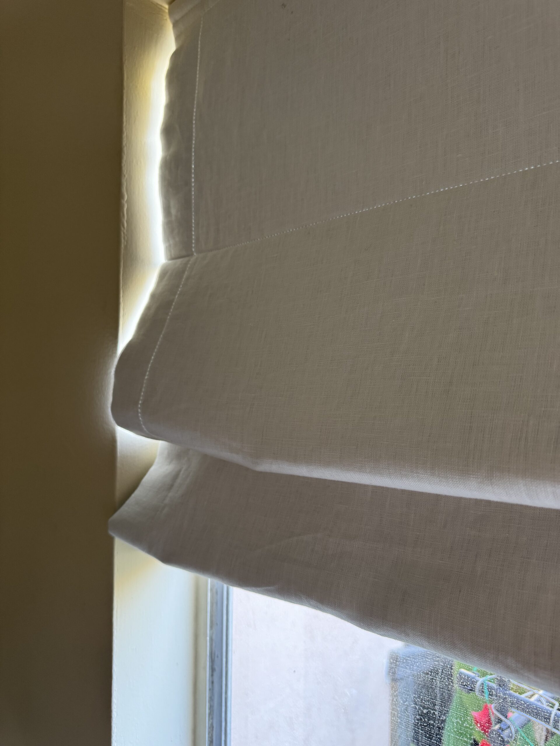 White fabric Roman shade partially pulled up near a window, with sunlight streaming through the sides.