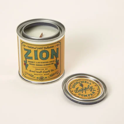 A candle in a tin container labeled "Zion" with the lid placed beside it; the label mentions desert lavender, sage, herbs, and sunrise.