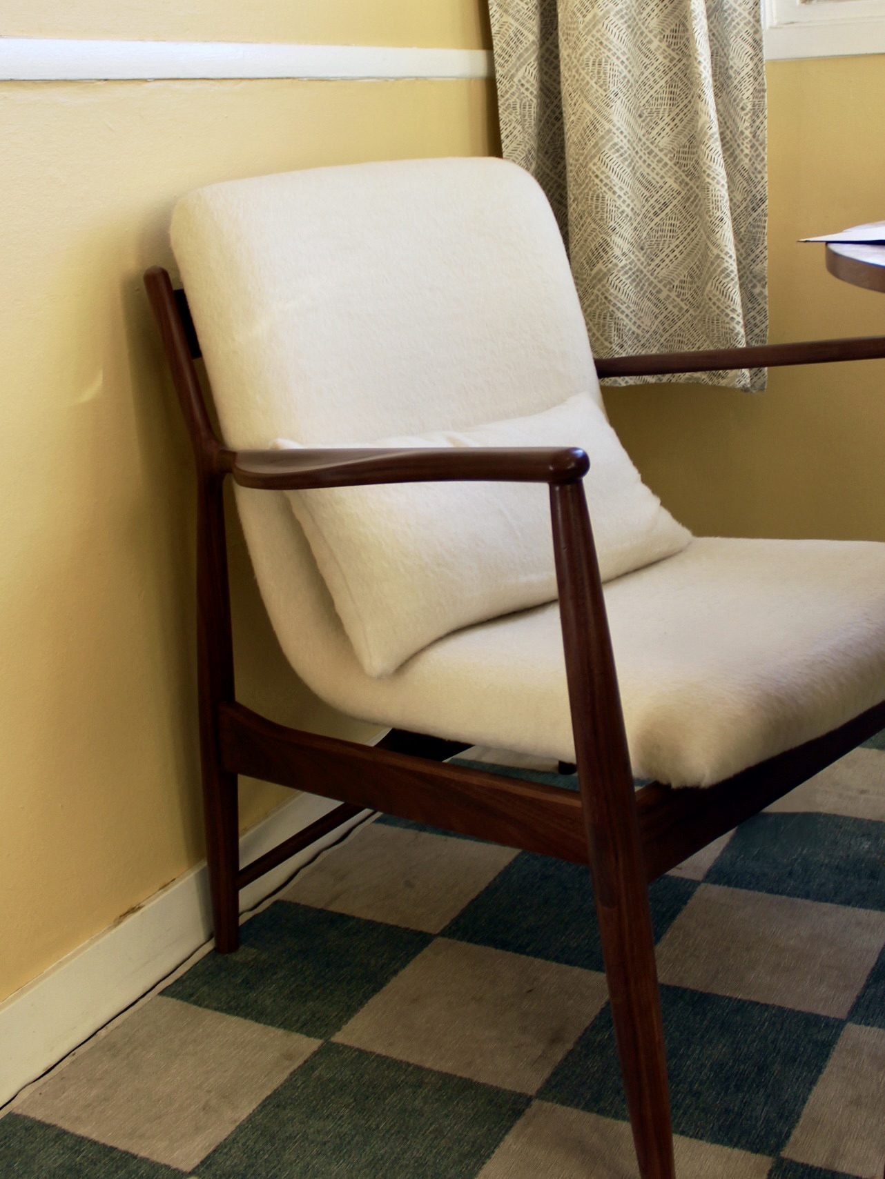 Mid-century modern chair with wooden arms and a cream cushion. Placed on a checkered rug near a beige wall and patterned curtain.