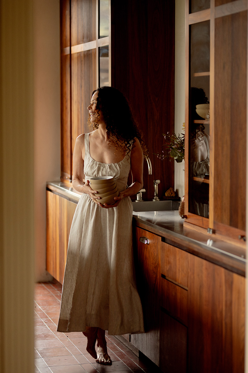 A woman in a long dress holds a bowl while standing in a warmly lit kitchen next to wooden cabinets.