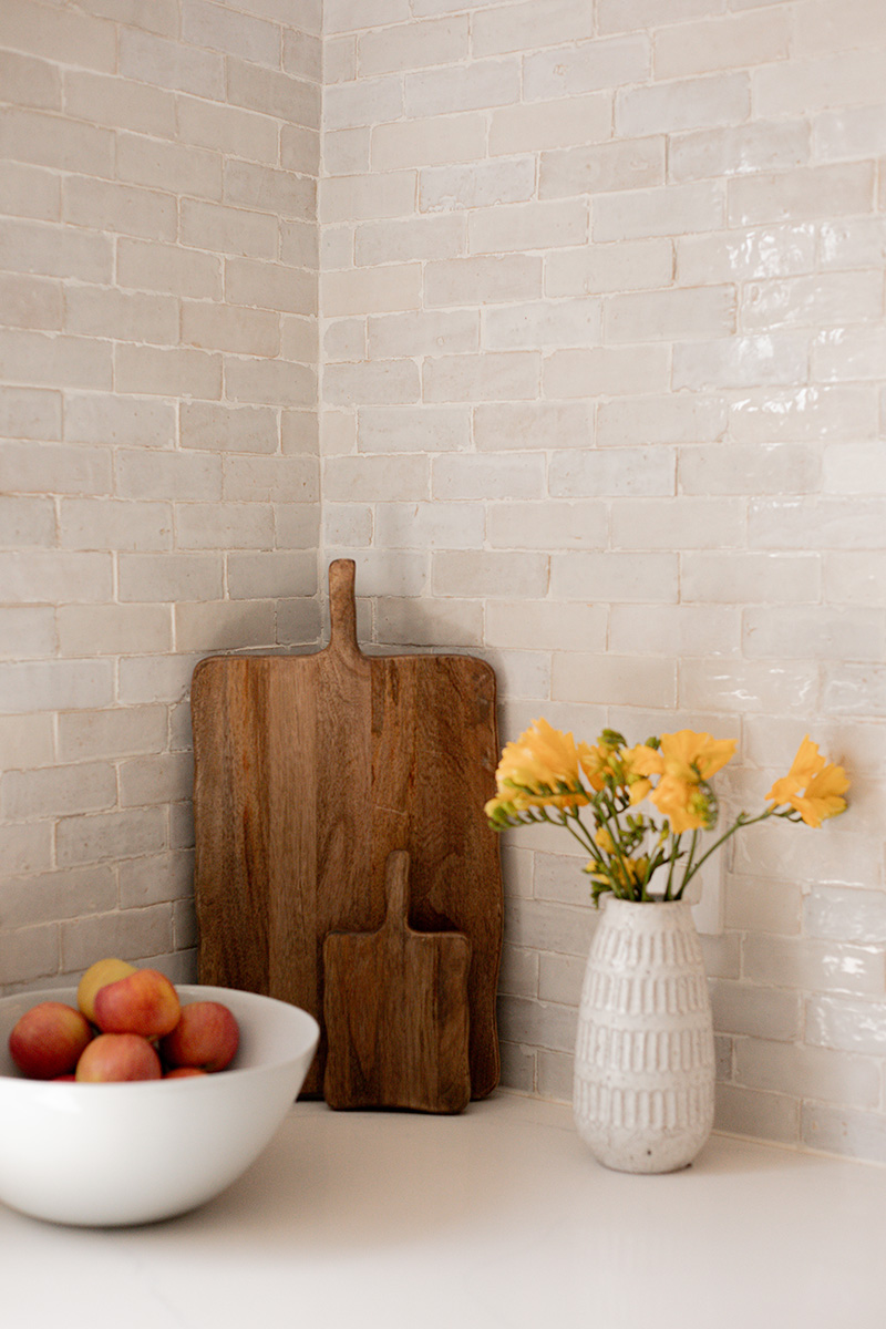 A kitchen corner with two wooden cutting boards, a vase with yellow flowers, and a bowl of apples on a white countertop against a tiled backsplash.