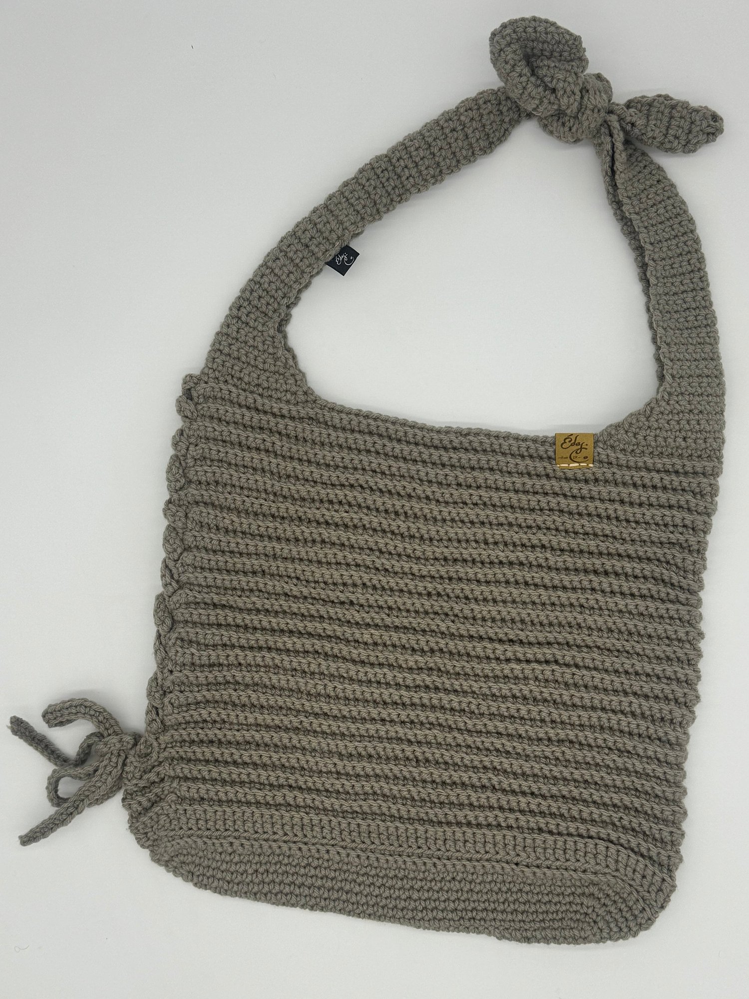 A handcrafted, knitted beige bag with braided detail, tied shoulder strap, and a small brown label near the top.