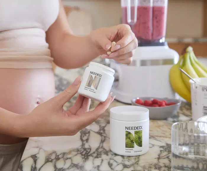 Pregnant person holding a supplement capsule and container near a marble countertop with a smoothie blender, bananas, and a bowl of raspberries in the background.