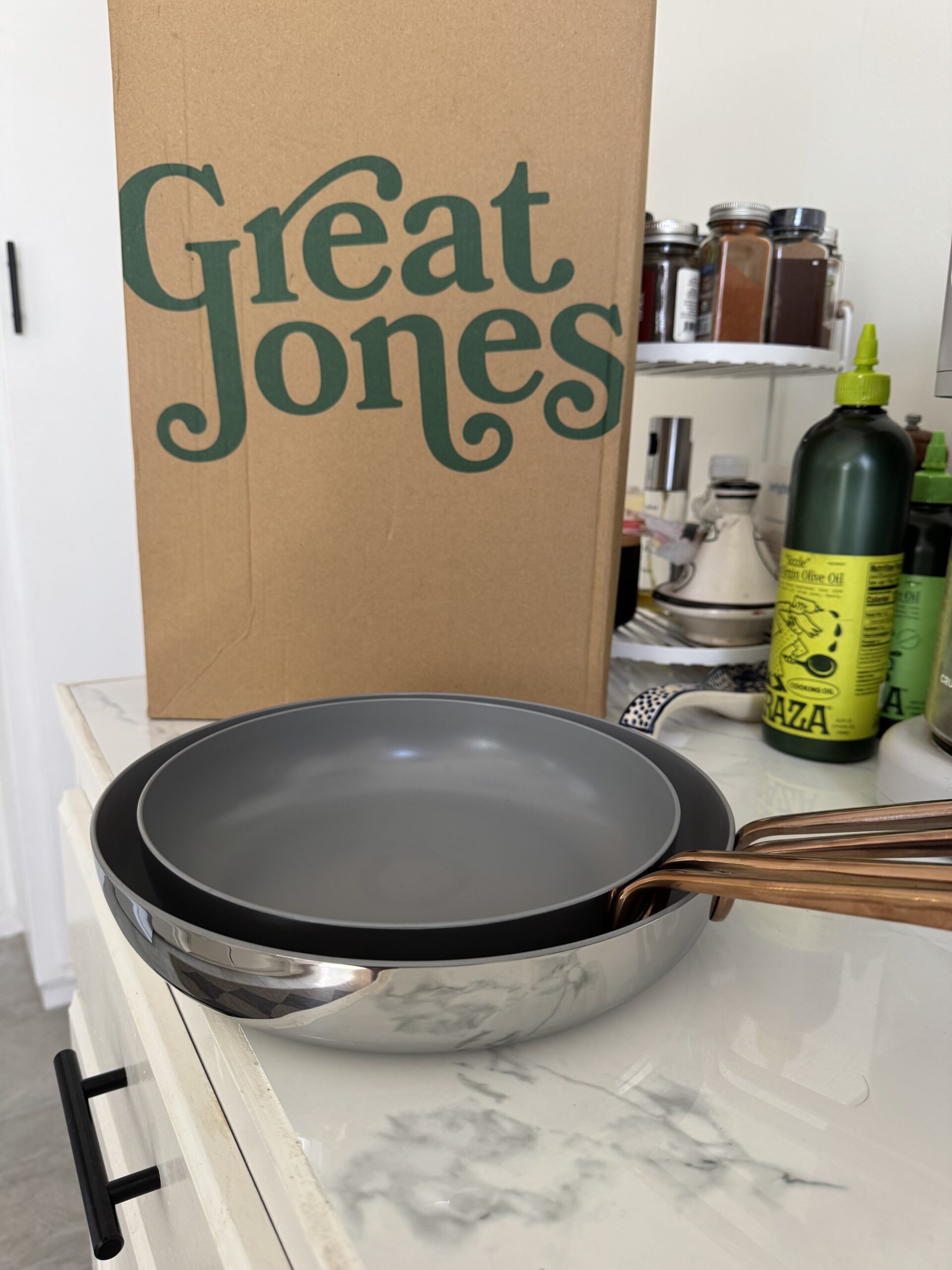 Two gray frying pans with wooden handles are stacked on a marble counter. A cardboard box with "Great Jones" and olive oil bottles are in the background.