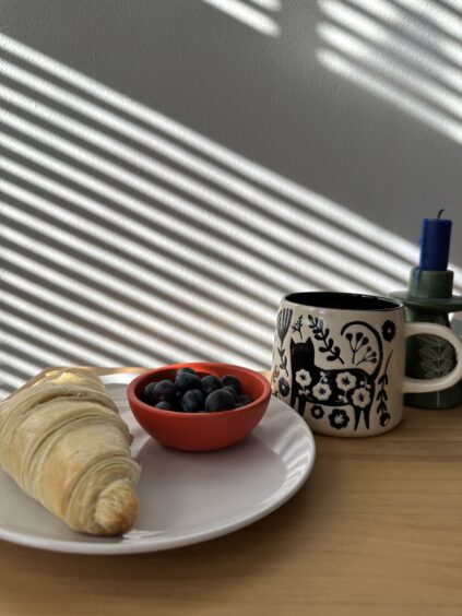 A croissant and bowl of blueberries on a white plate beside a patterned mug with a candle, on a wooden table with striped sunlight.
