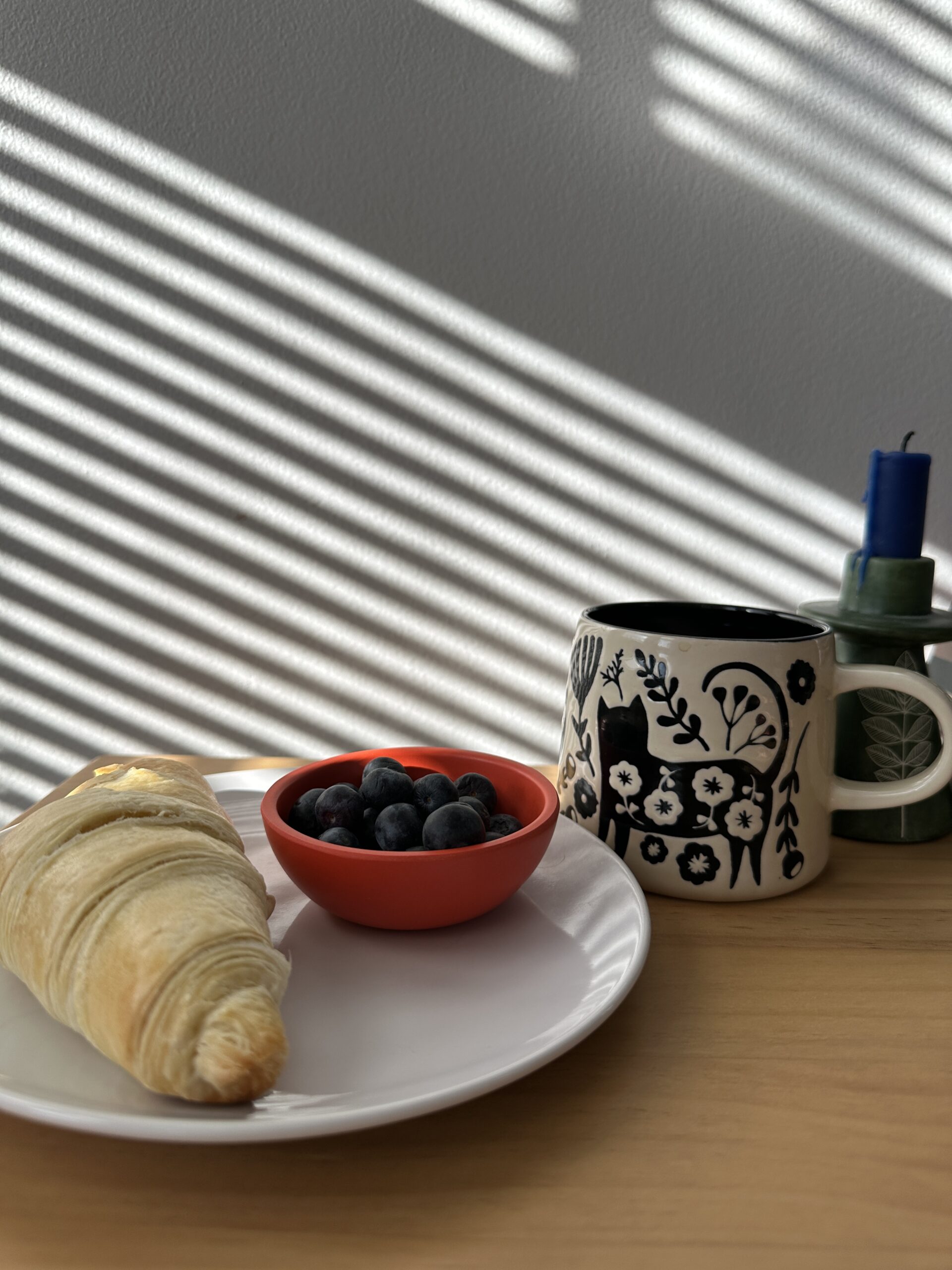 A croissant and bowl of blueberries on a white plate beside a patterned mug with a candle, on a wooden table with striped sunlight.