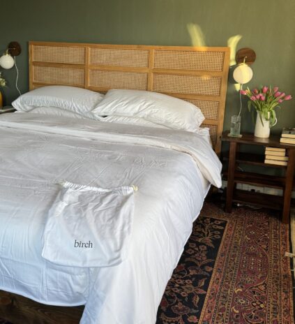 Bedroom with a double bed, white linens, and a rattan headboard. There are two bedside tables, flowers, a yellow vase, and a patterned rug.