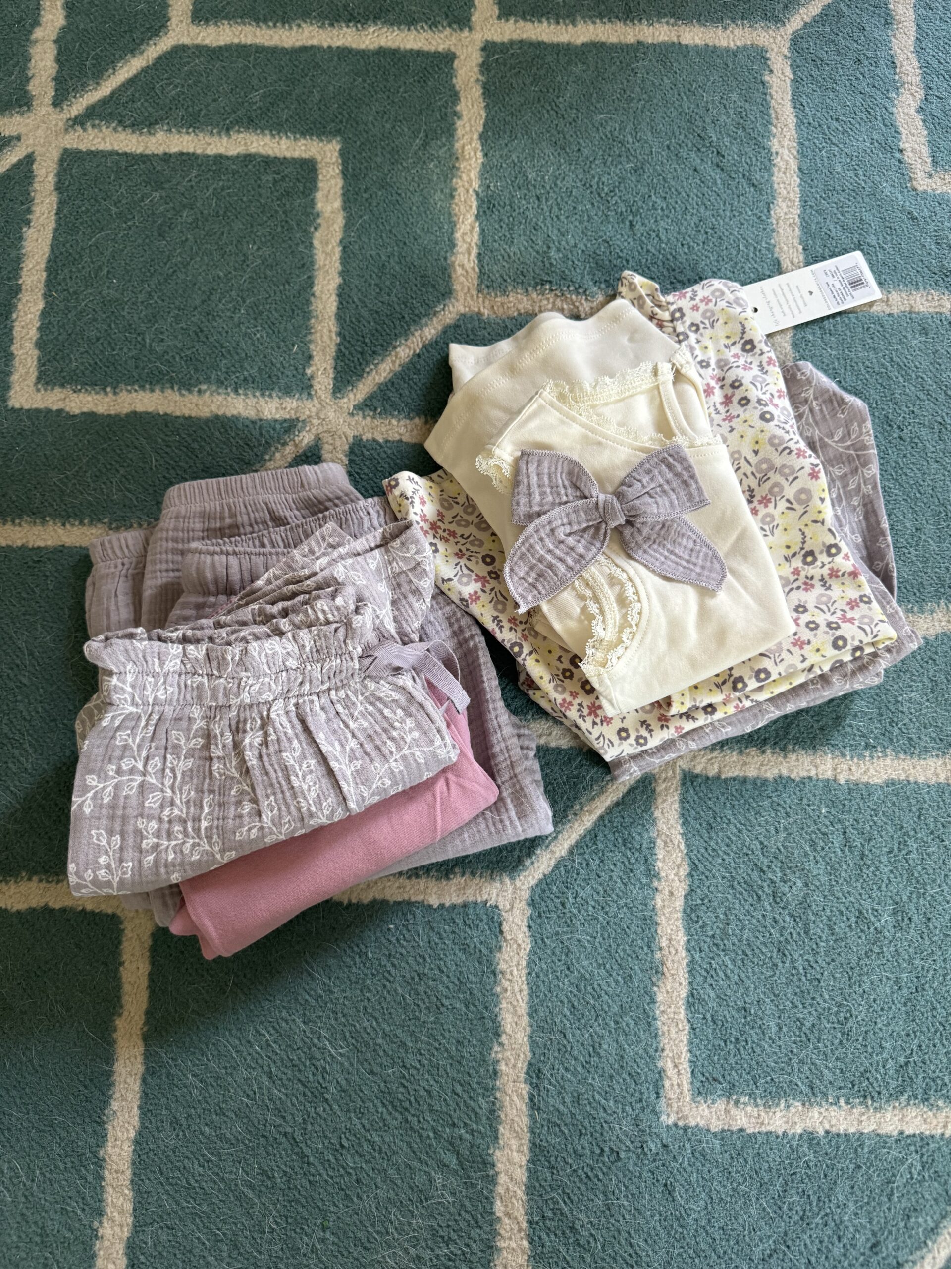 Two piles of folded children's clothing on a green patterned rug; one pile is mostly pink and purple, the other contains floral and cream items, some with lace and a bow.