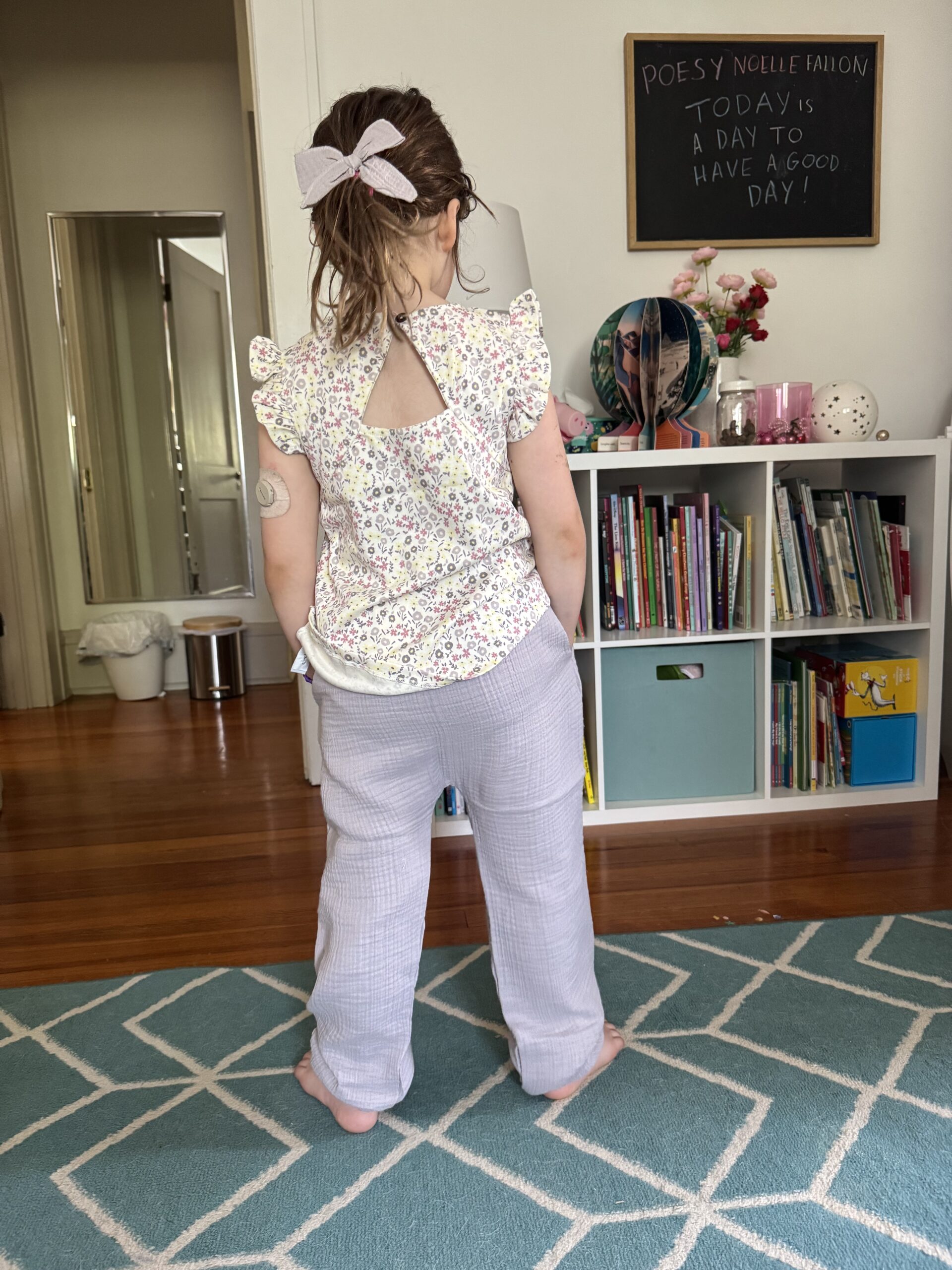 A young child with a bow in their hair stands barefoot on a blue rug, facing away, in a room with a bookshelf, a mirror, and a chalkboard with writing.