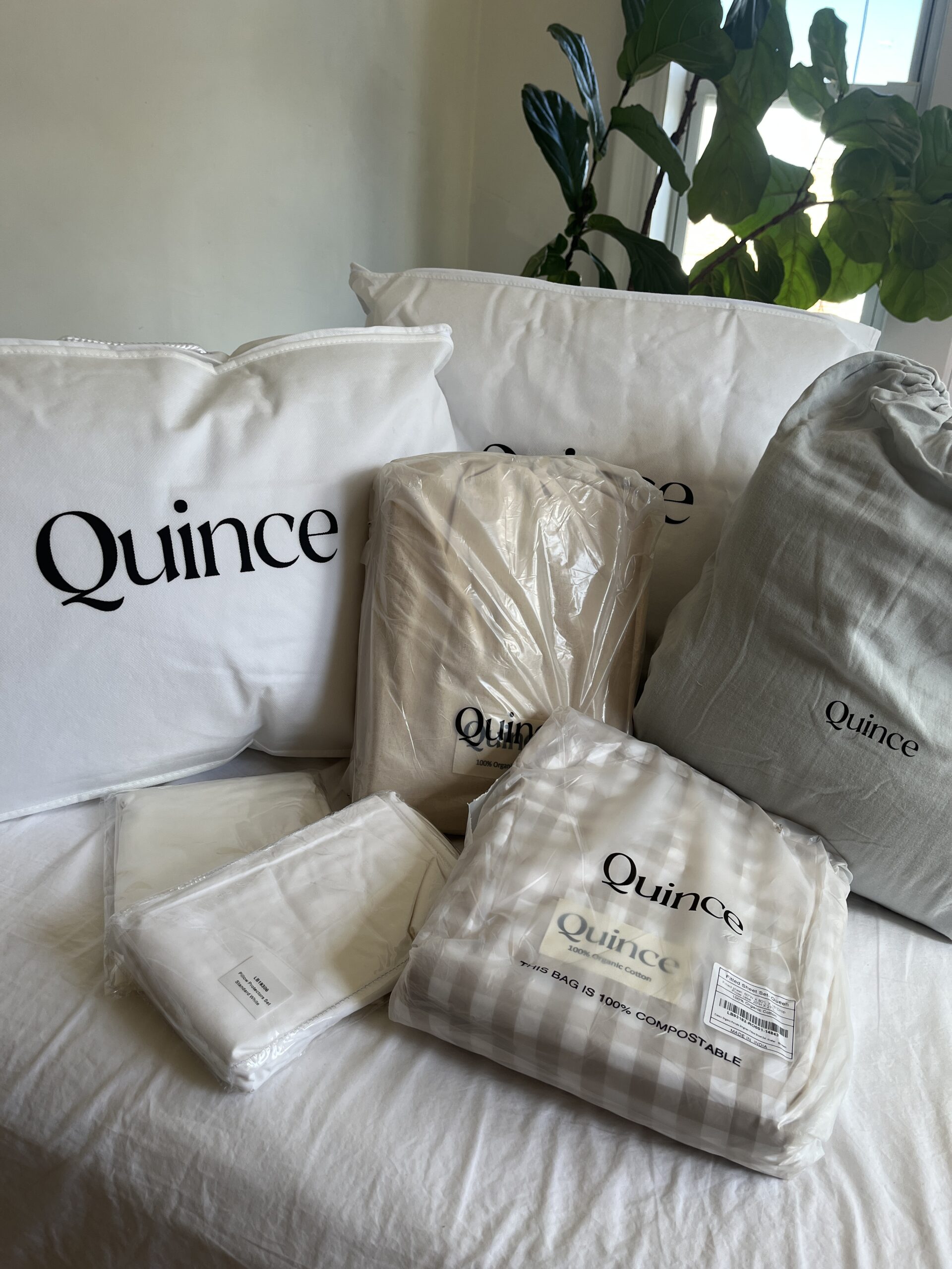 Quince branded bedding items, including pillows, blankets, and sheet sets, arranged on a bed. A leafy plant is visible in the background.