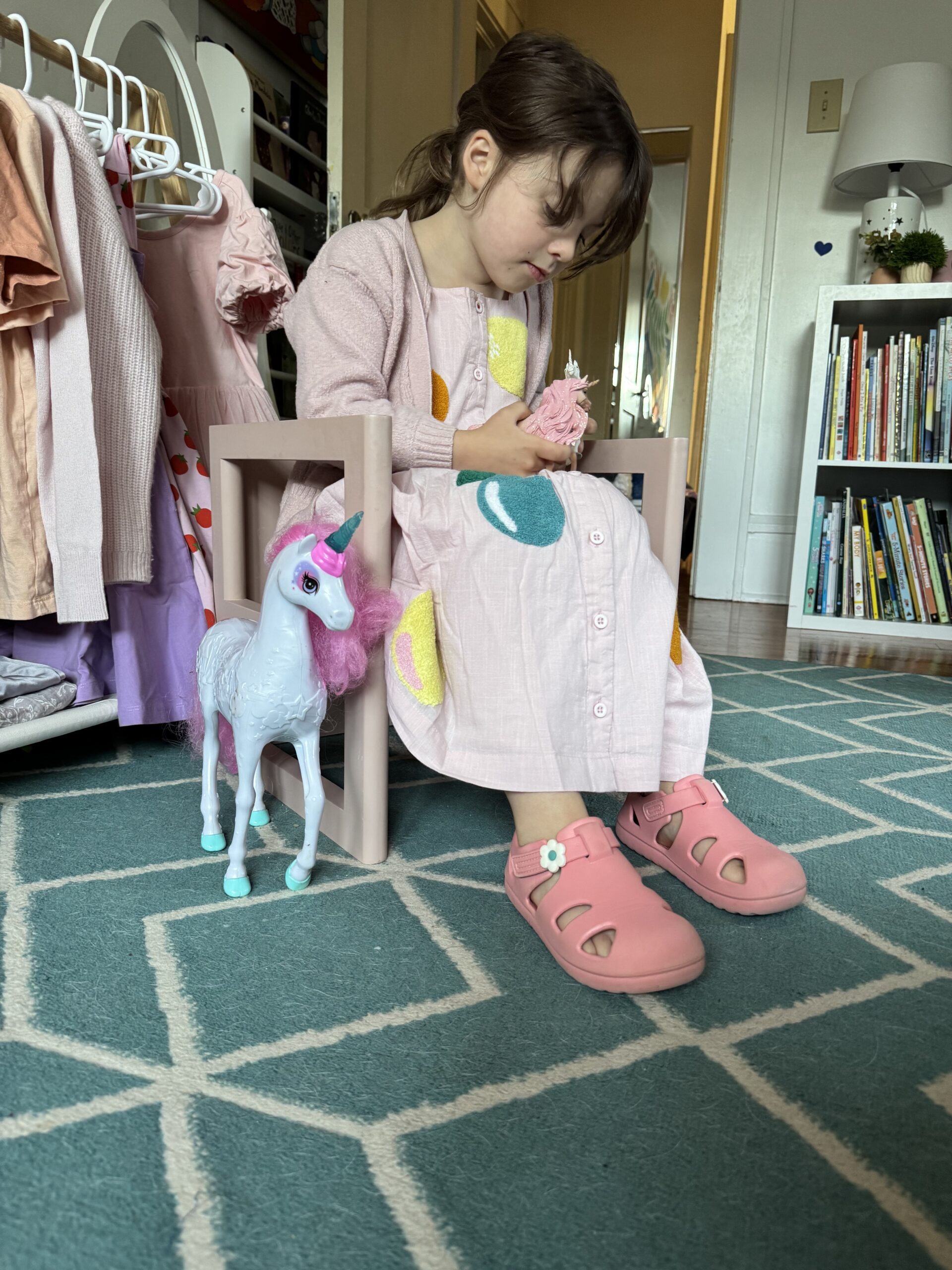 A young girl in a pink dress and shoes sits on a small chair holding a toy, with a unicorn figurine on the floor beside her in a room with bookshelves and clothes.
