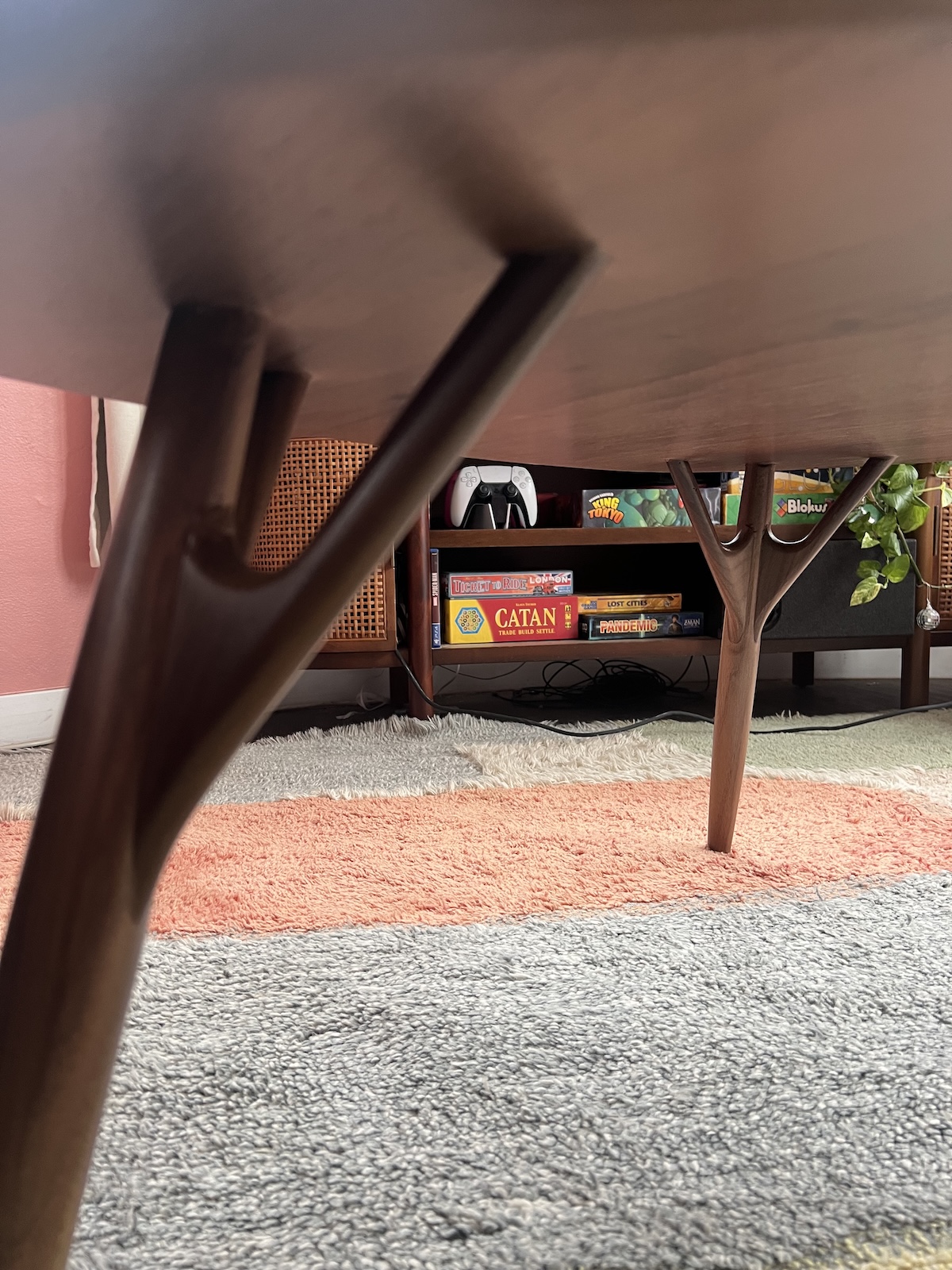 View from under a wooden coffee table showing board games on a shelf, including Catan, against a backdrop of pink and gray carpets.