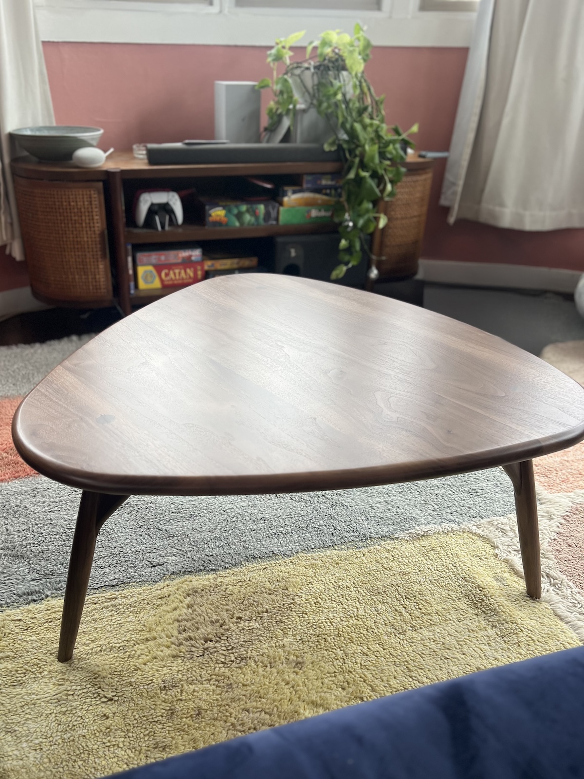 A wooden triangular coffee table on a layered rug in front of a wooden shelf with plants, books, and speakers.