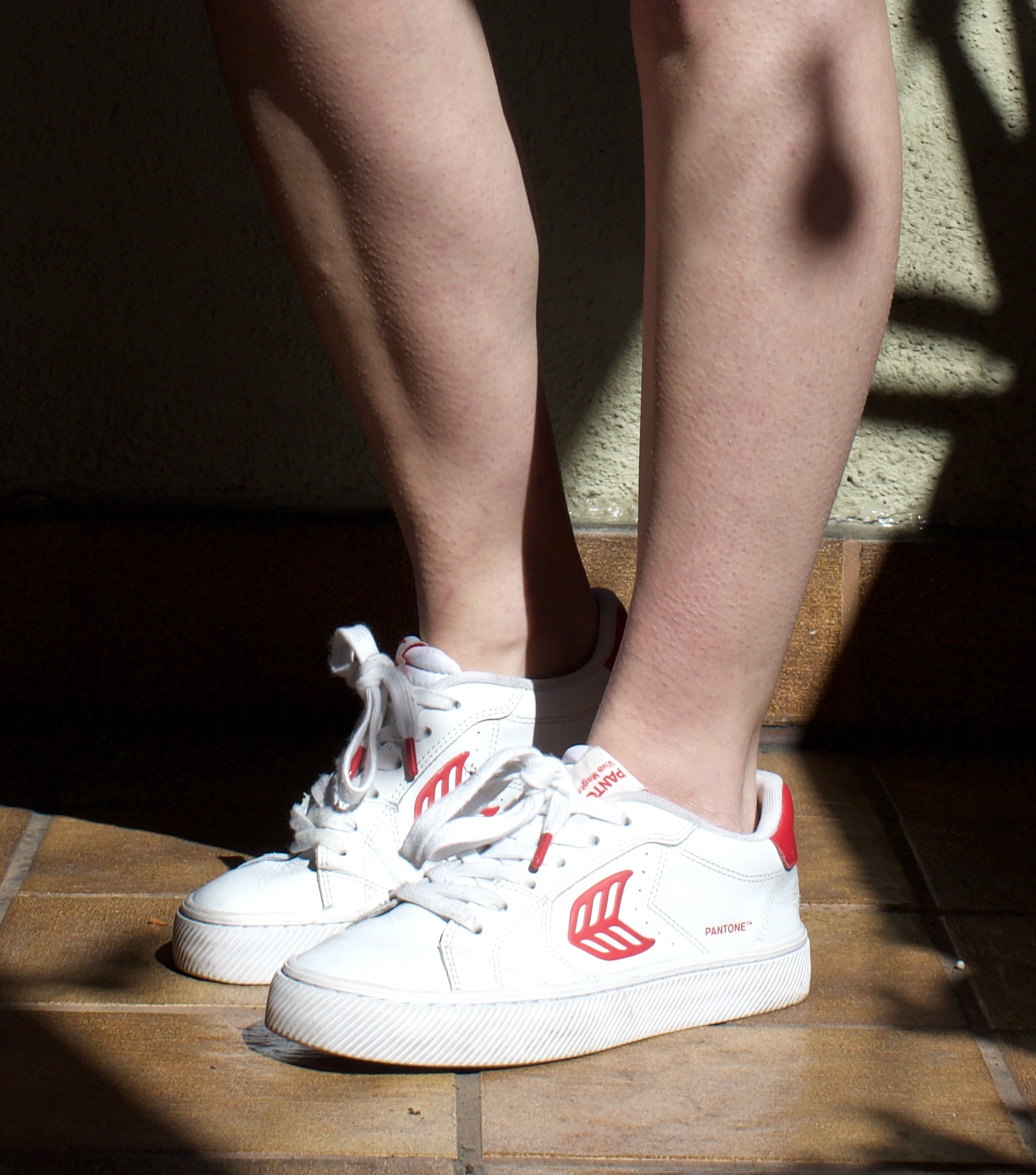 A person stands on a tiled floor wearing sustainable sneakers—white with red accents and white laces—as sunlight casts soft shadows on their legs.