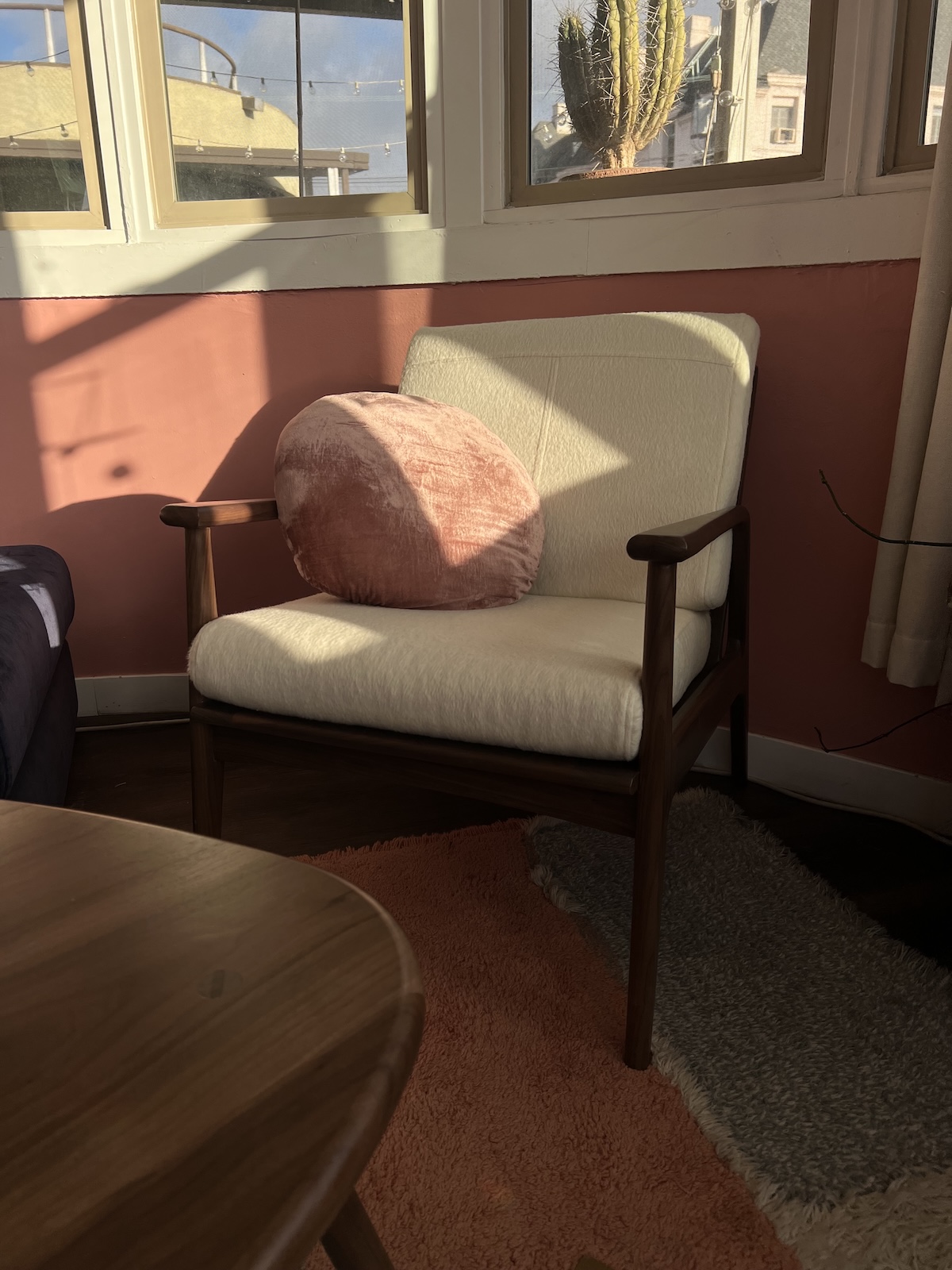 A white cushioned chair with a round pink pillow is placed next to a window in a sunlit room with pink walls.