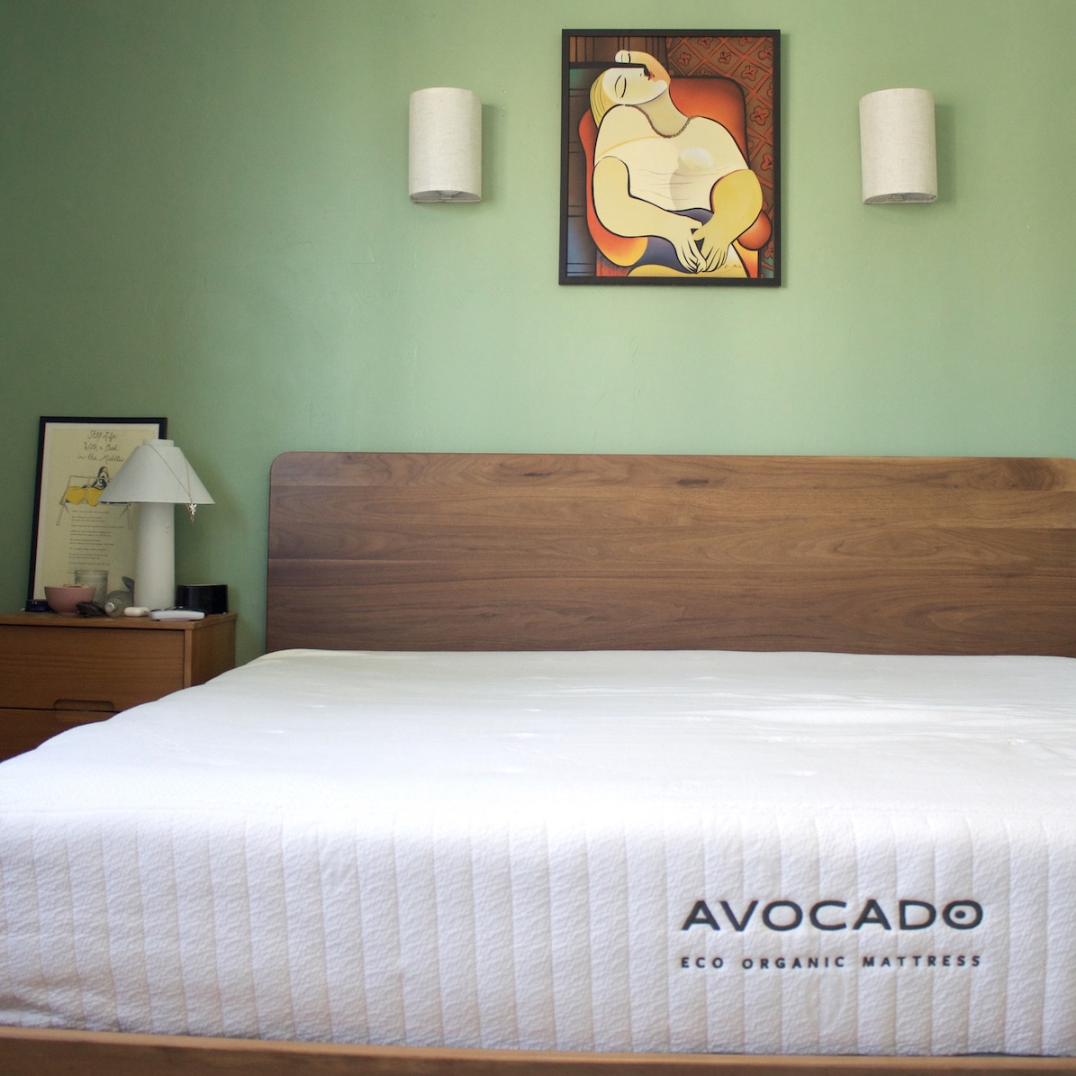 Bedroom with a wooden bed frame and white mattress labeled "Avocado Eco Organic Mattress." Light green wall with abstract art and two white wall lamps.