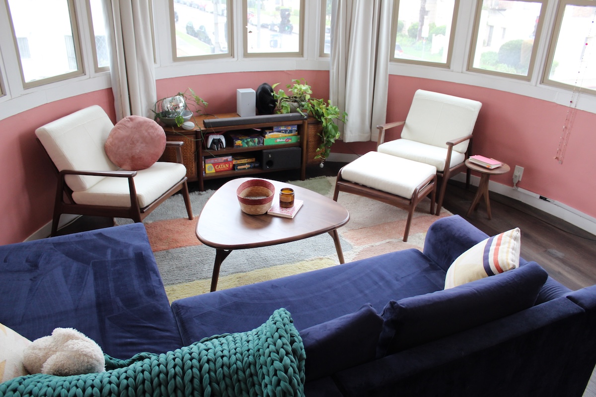 A cozy living room with a navy sectional sofa, two white armchairs, a wooden coffee table, and a bookshelf. The walls are pink, and large windows provide natural light.