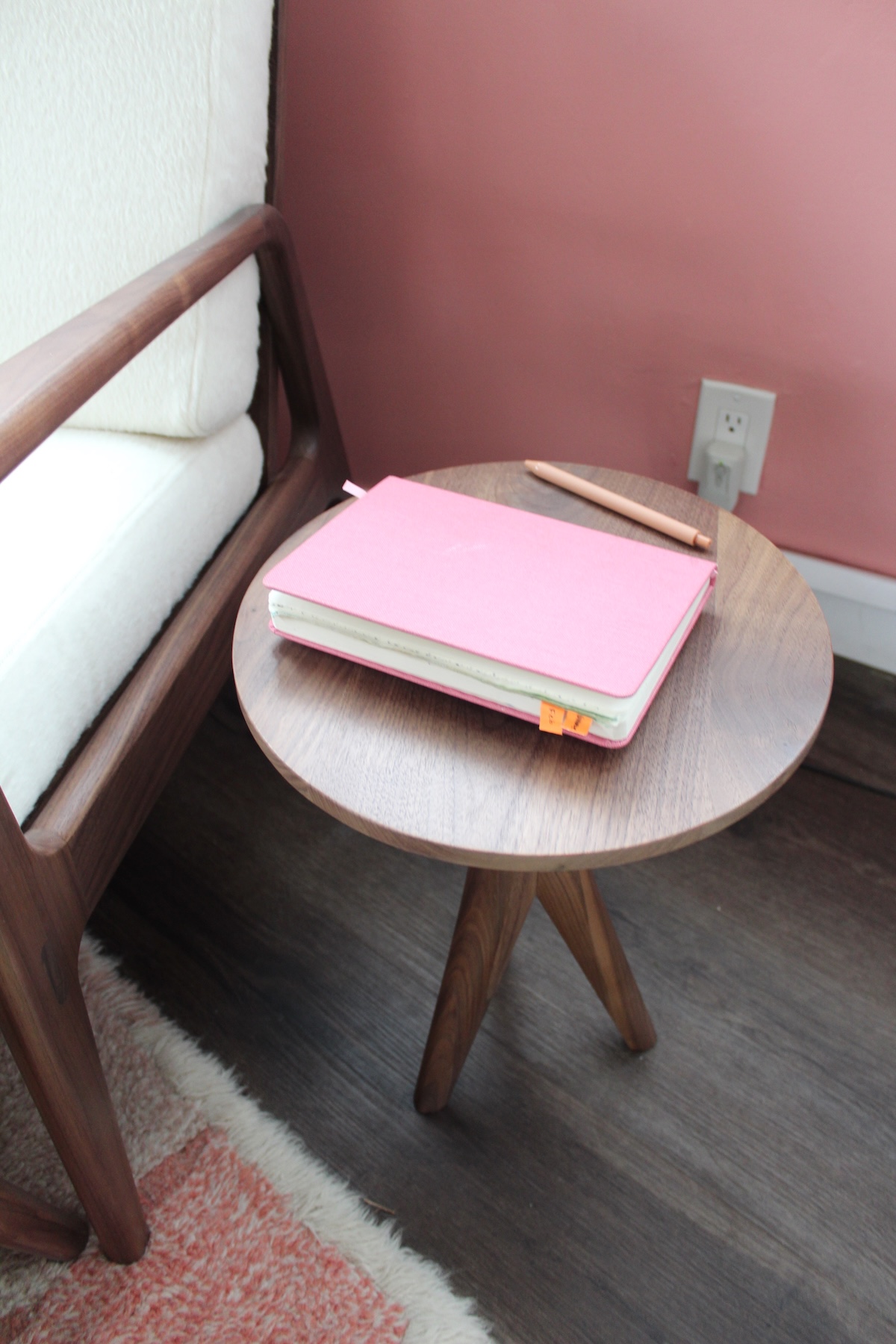 A small round wooden table with a pink notebook and a pen on top. The table is next to a cushioned chair, set against a pink wall.