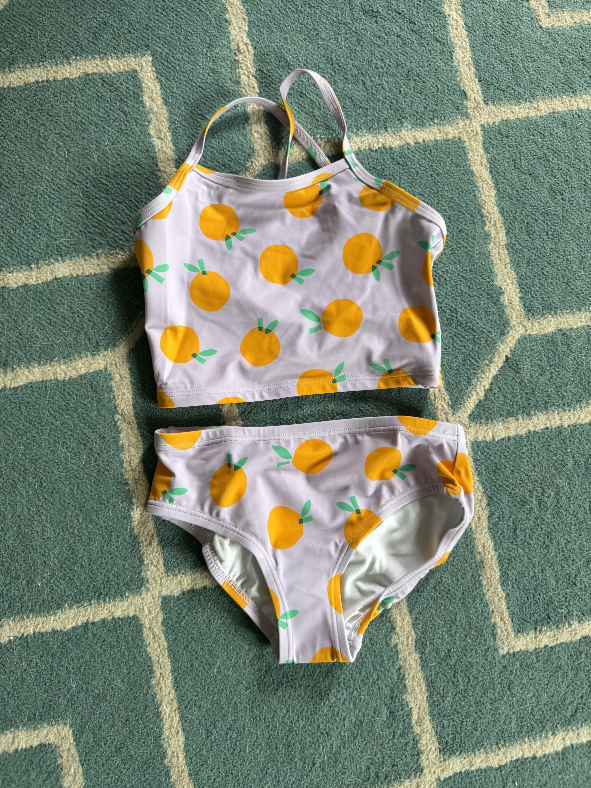 A two-piece swimsuit with an orange fruit print is laid flat on a green and white geometric-patterned rug.