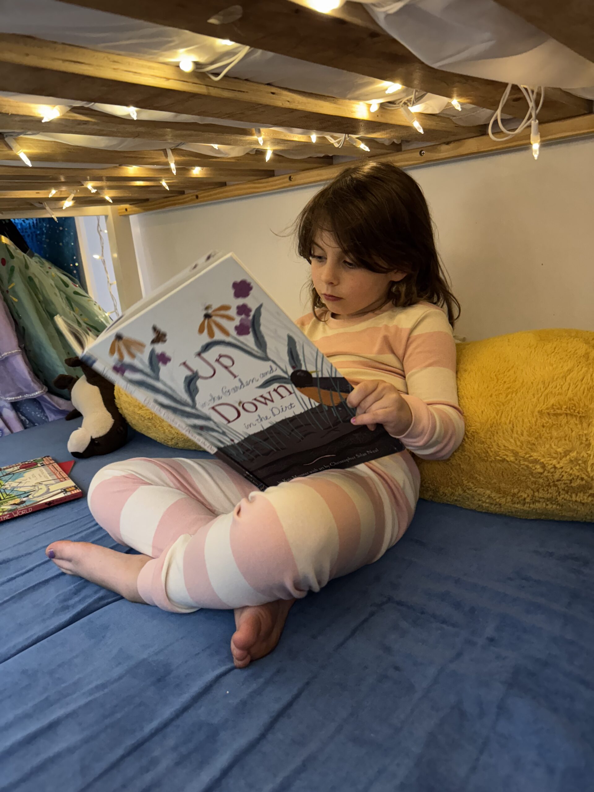 A young child in striped pajamas sits on a blue bed with a yellow pillow, reading a picture book under warm string lights.